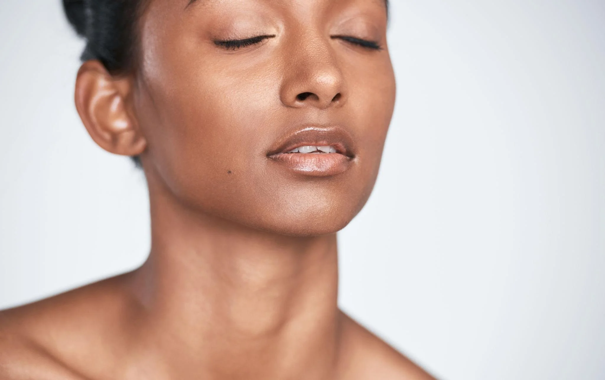 Close-up of a woman's face with closed eyes, warm skin tone, and natural makeup, against a plain light background.