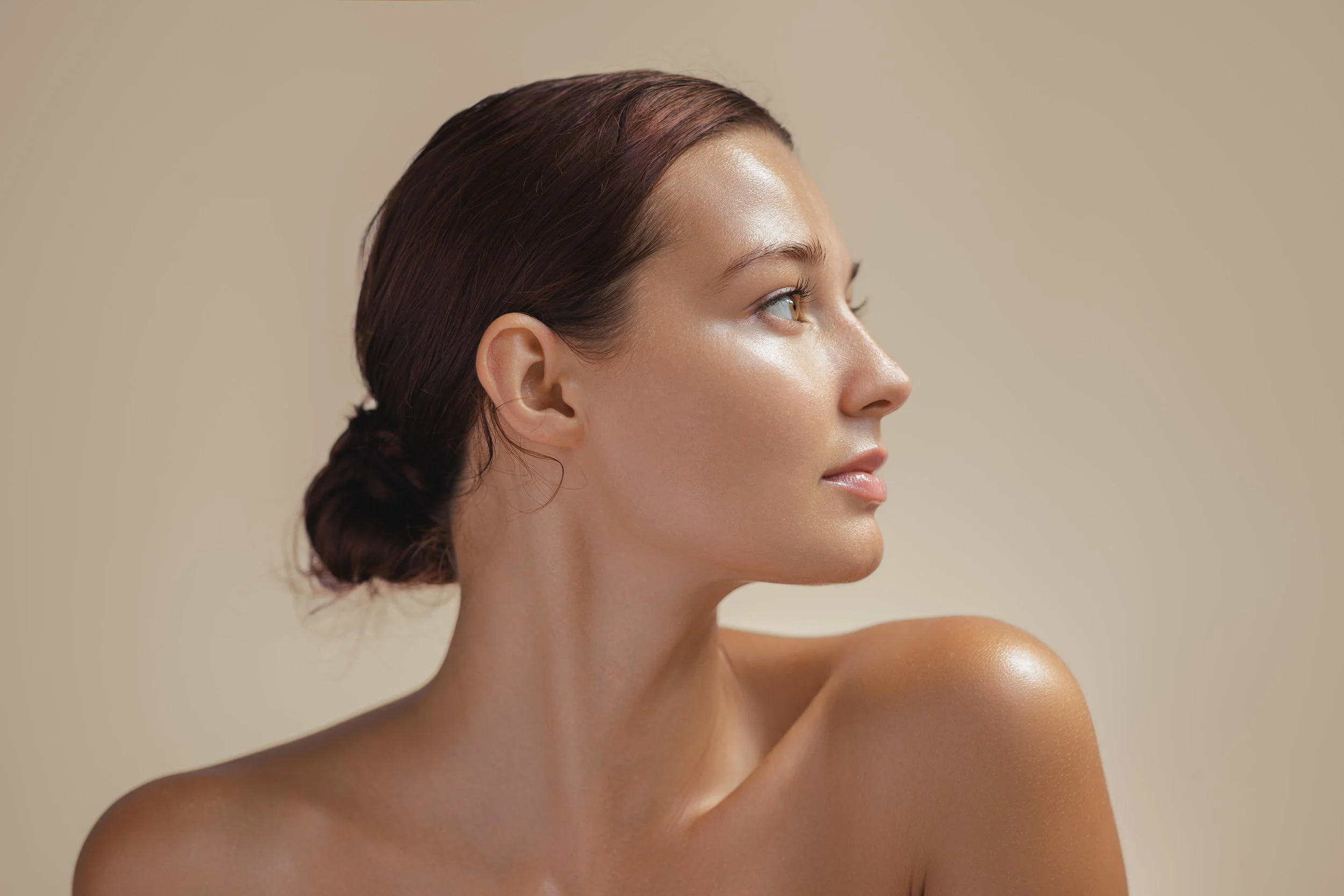 Profile of a woman with smooth skin, brown hair tied back, looking to her right, against a neutral beige background.