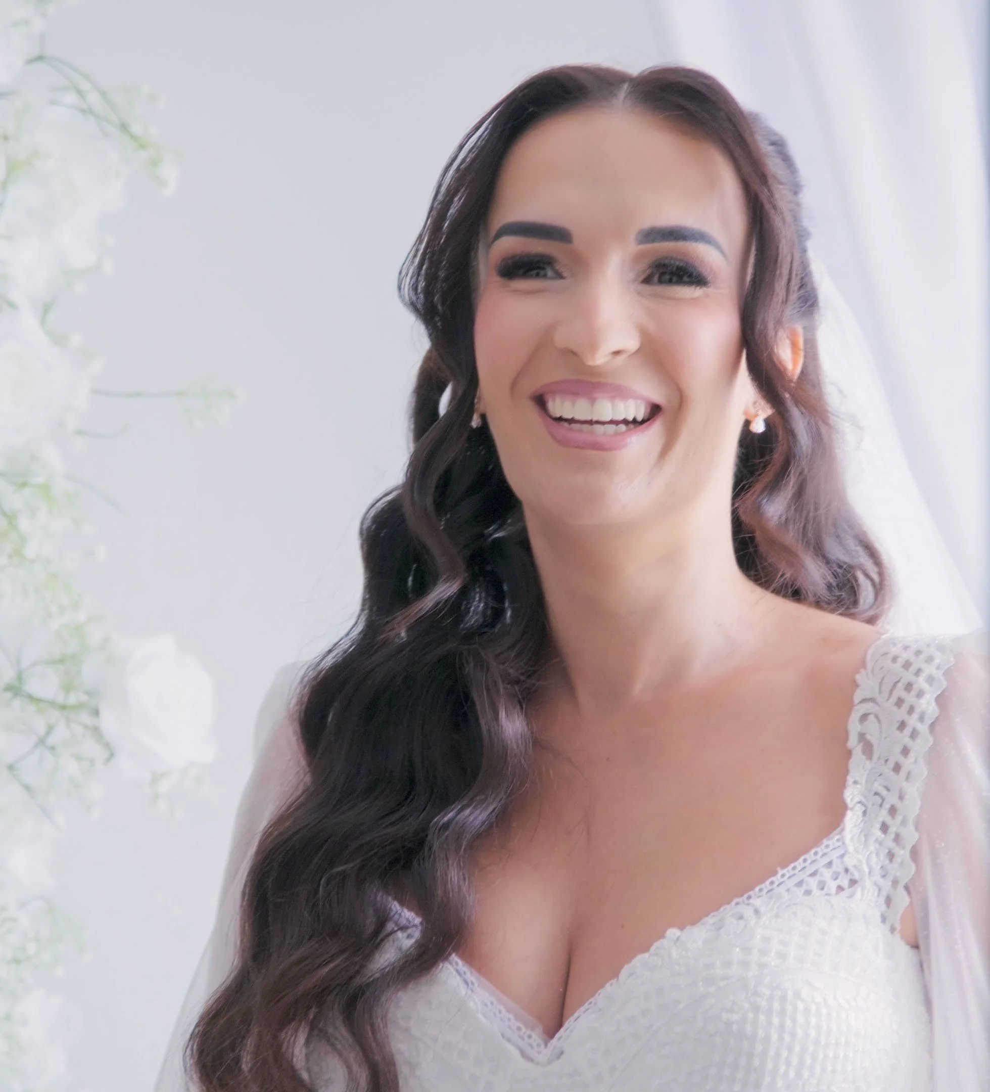 A woman with long dark hair smiling and wearing a white lace dress, near a white curtain and light-colored background.