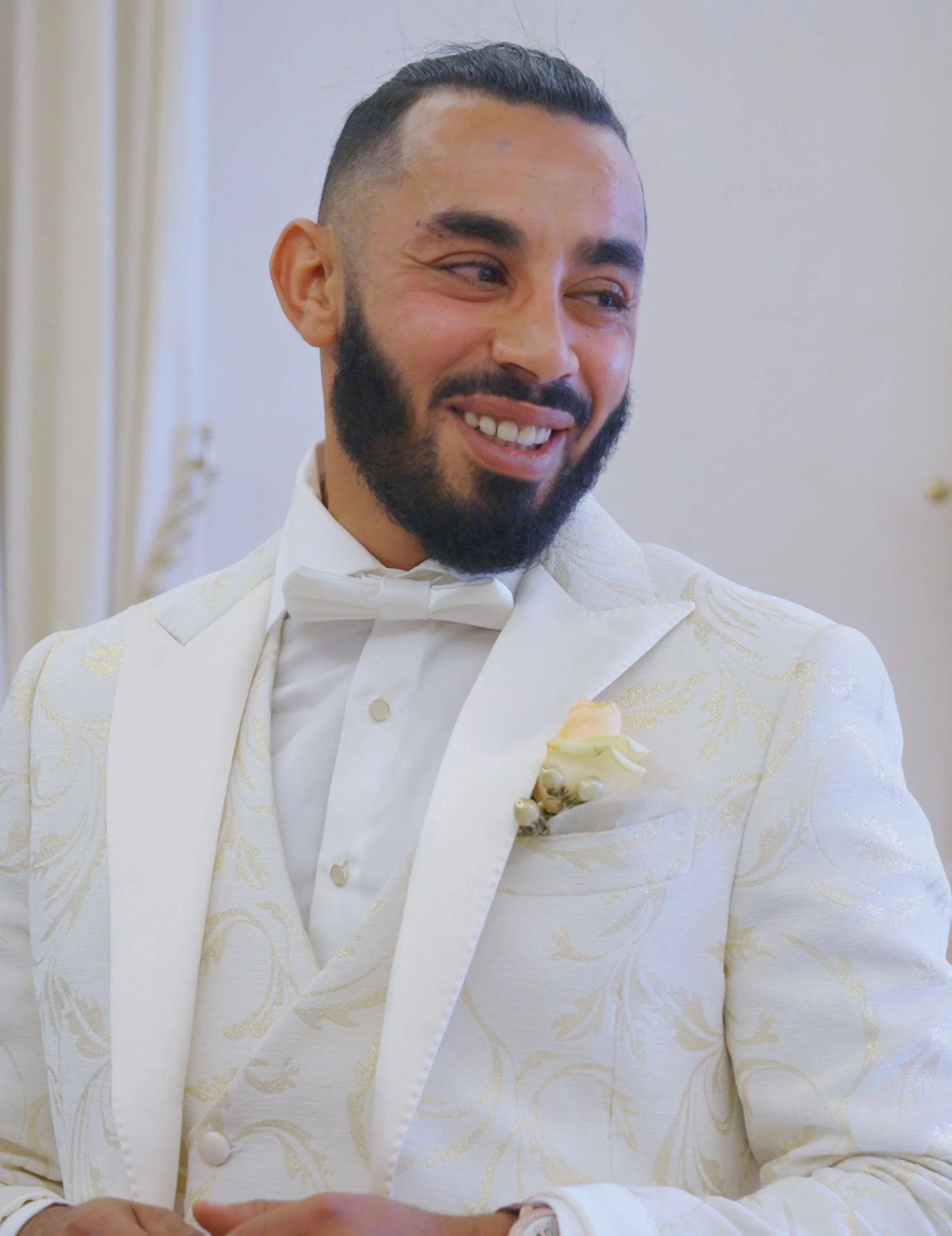 A man dressed in a white suit with gold accents and a white bow tie, smiling with a visible smile line, in a light-colored room with a neutral background.