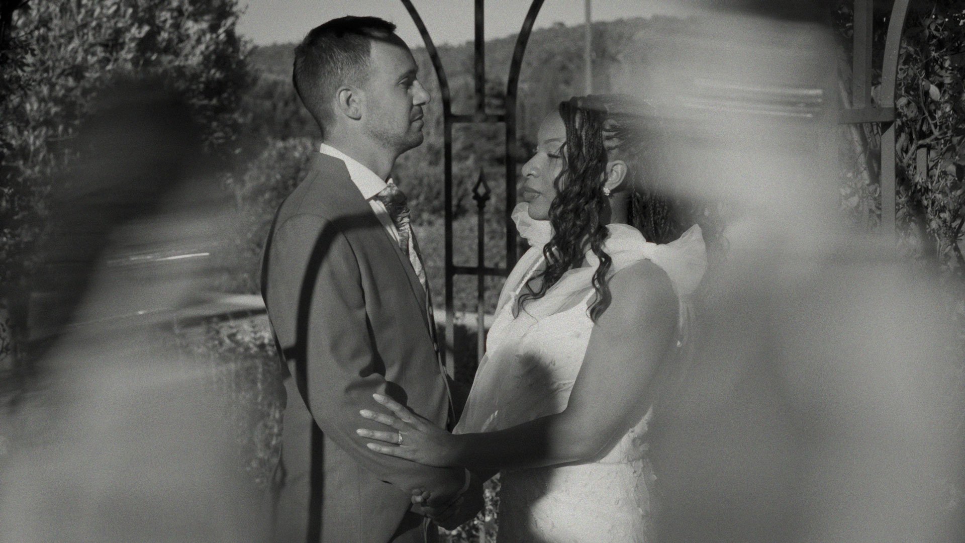 A black and white photograph of a couple standing close together outdoors, possibly at a wedding or special event. The man is in a suit, and the woman is in a dress. They are facing each other, holding hands, with trees and decorative ironwork in the background.