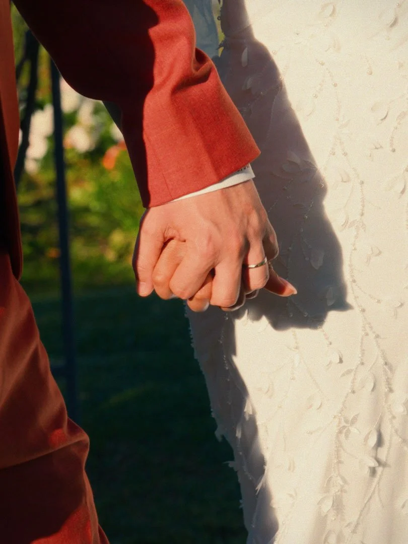 Close-up of two hands holding each other, one hand with a wedding ring, against a light-colored fabric background with a floral pattern.