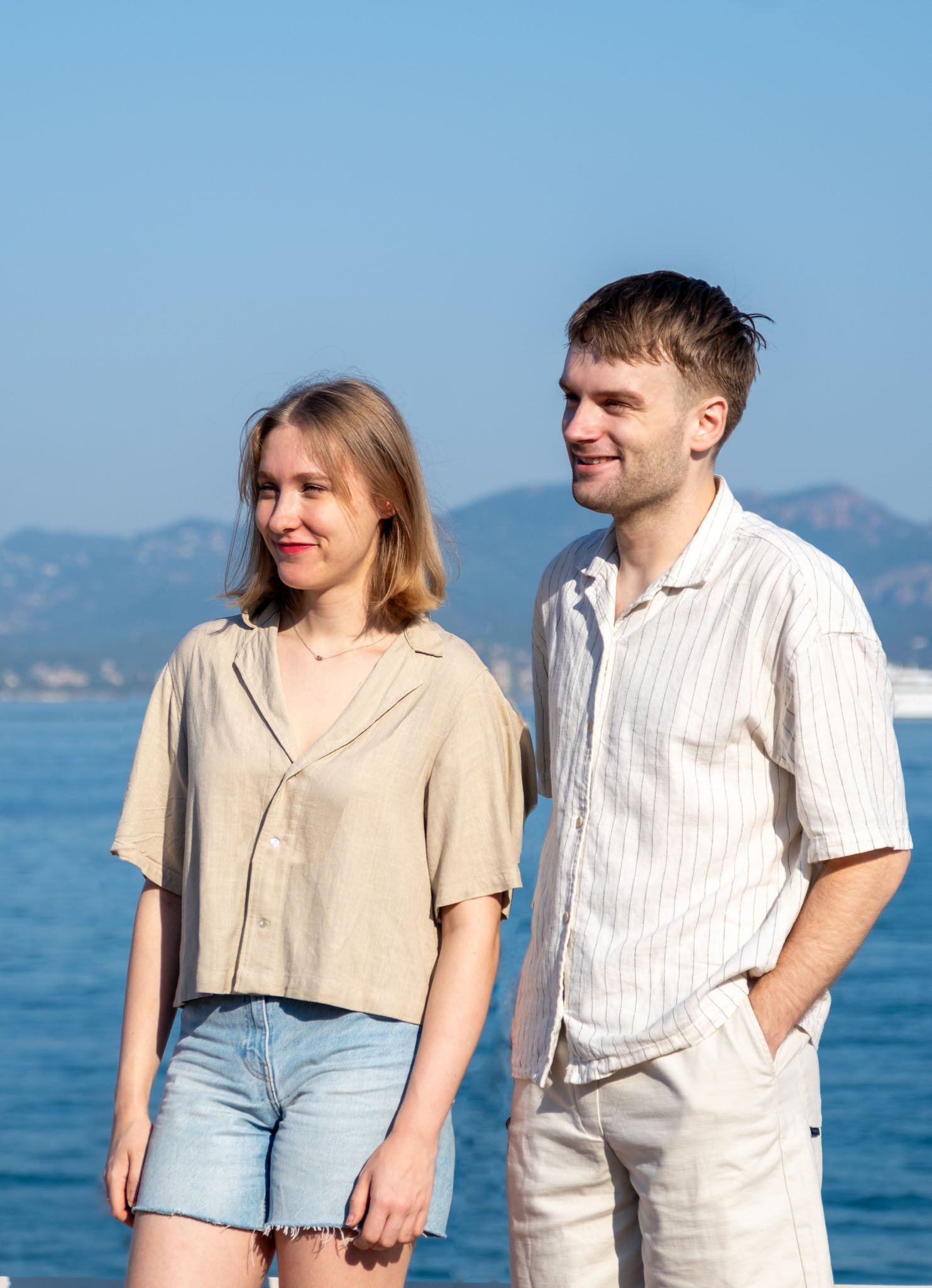 A young woman and a young man standing outdoors near water, with mountains in the background, smiling and enjoying a sunny day.