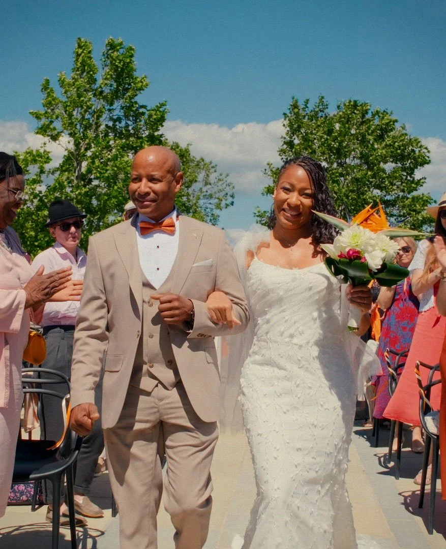 Bride walking down the aisle with her father, holding a bouquet of flowers, surrounded by guests at an outdoor wedding.