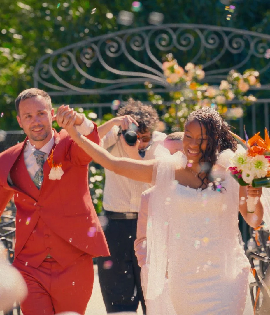 A newlywed couple celebrating outdoors, with the groom in a red suit and the bride in a white dress holding a bouquet, as they walk together amid confetti and a photographer capturing the moment.