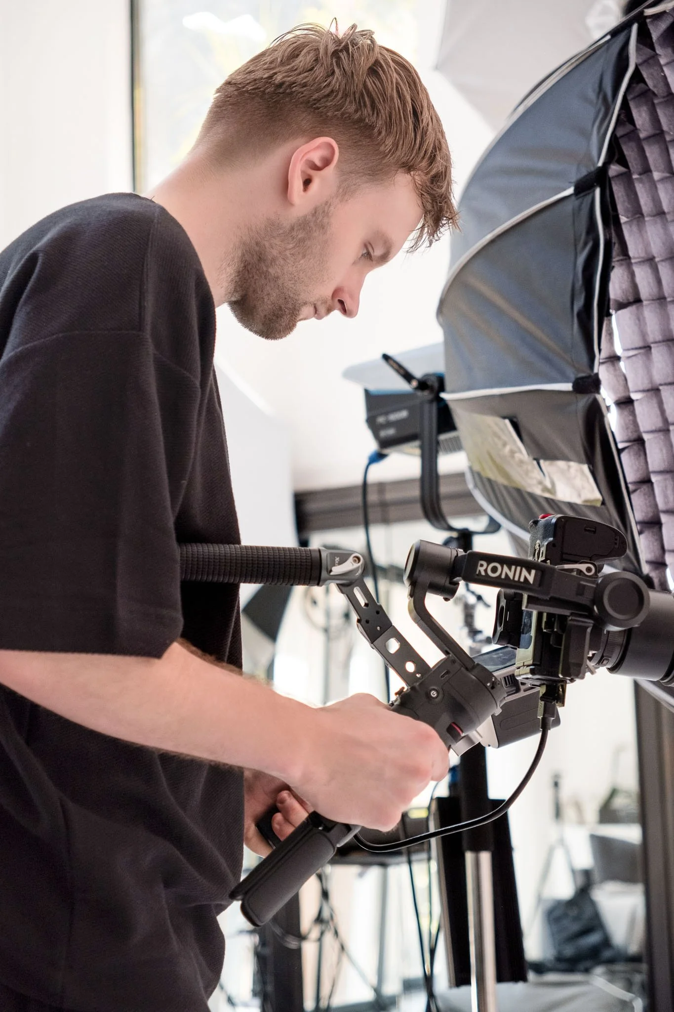 A man wearing a black shirt working with a professional camera mounted on a tripod, indoors near a window with bright natural light.
