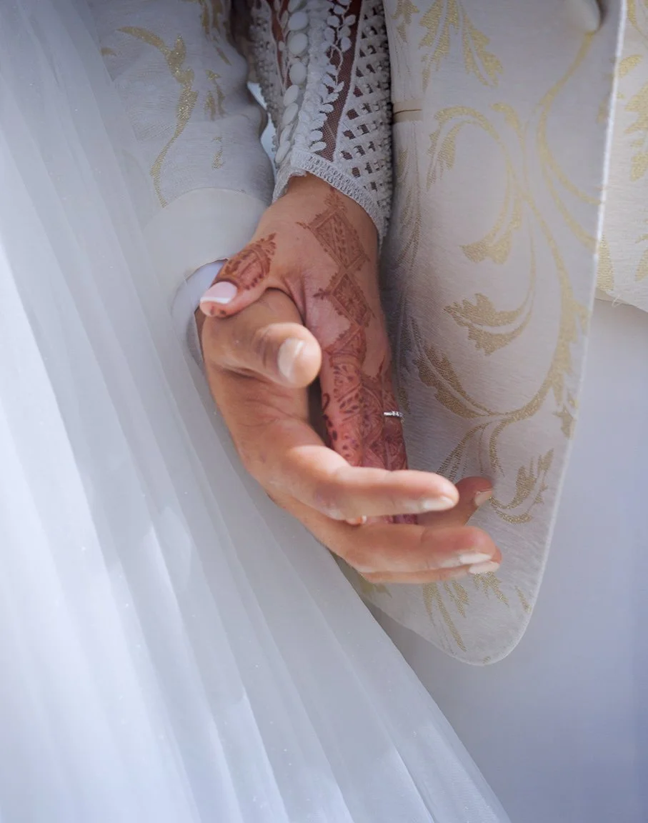 Close-up of a woman's hand with intricate henna (mehndi) designs, resting on a surface with gold-patterned fabric and sheer white material in the background.
