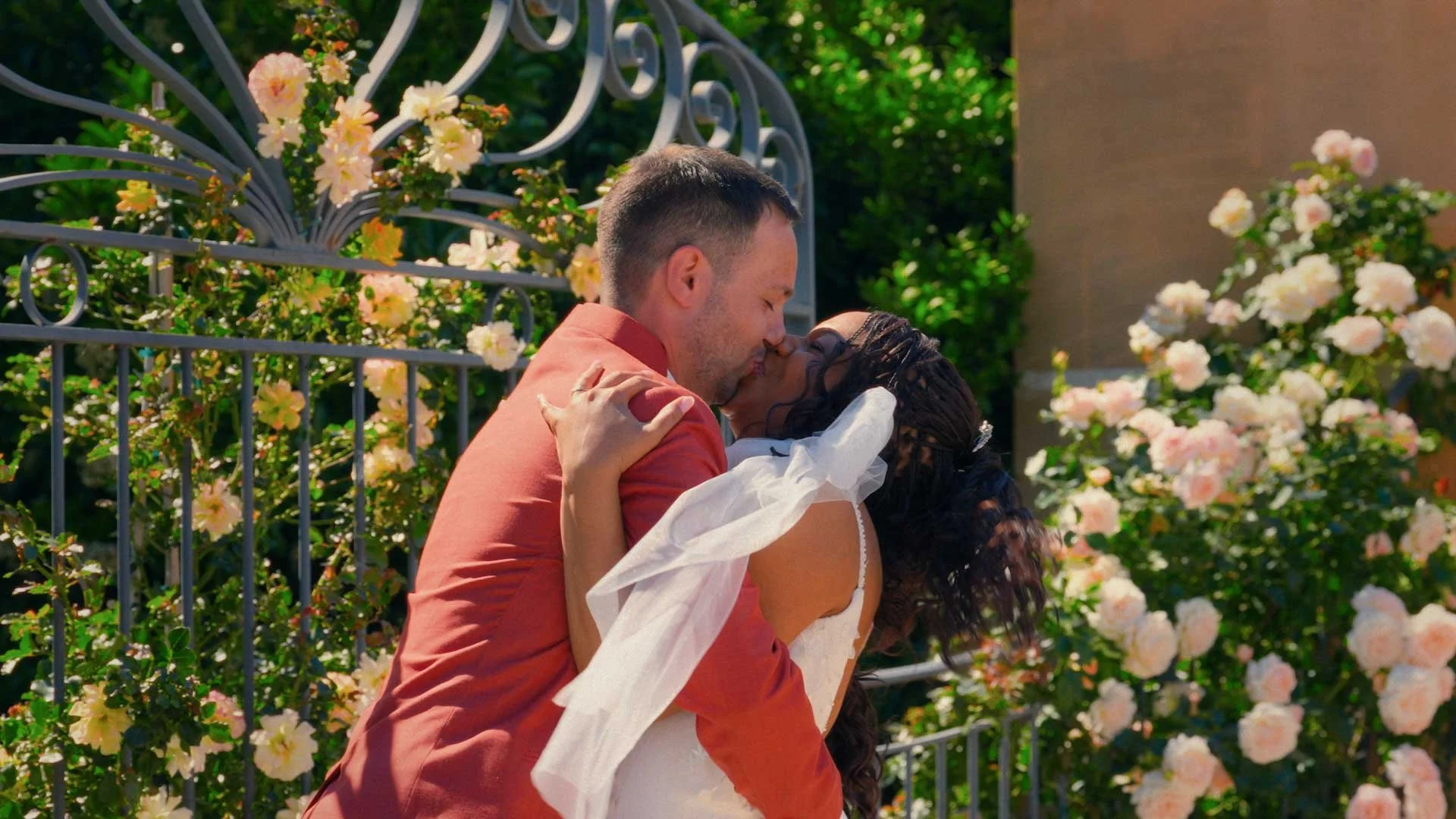 A couple sharing a kiss outdoors near a black metal gate with pink and white roses in the background.