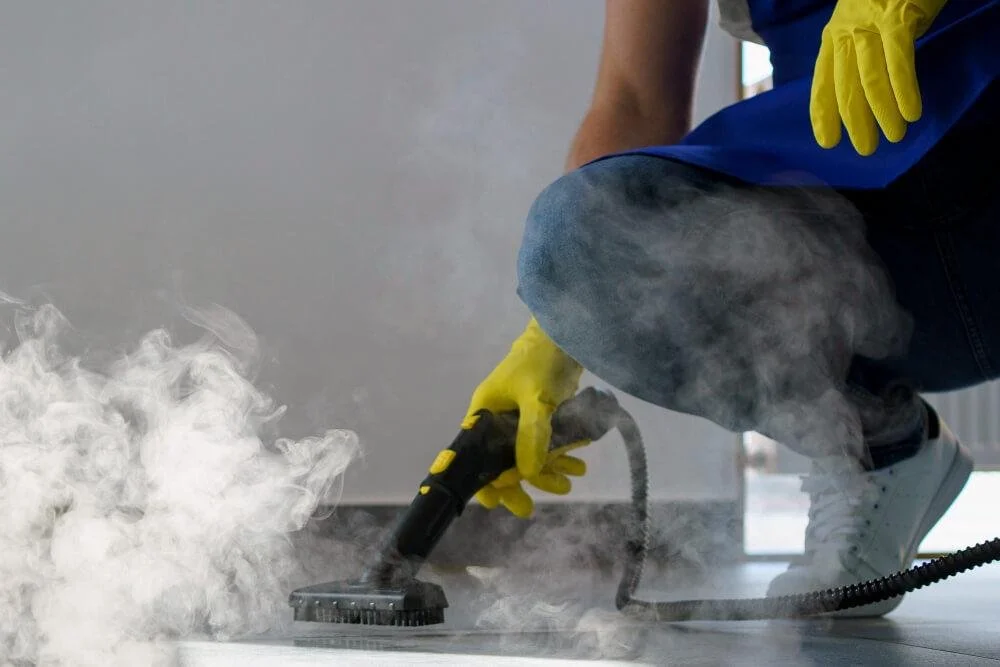 Person using a steam cleaner on the floor wearing yellow gloves, blue shirt, jeans, and white sneakers, with visible steam and haze around.