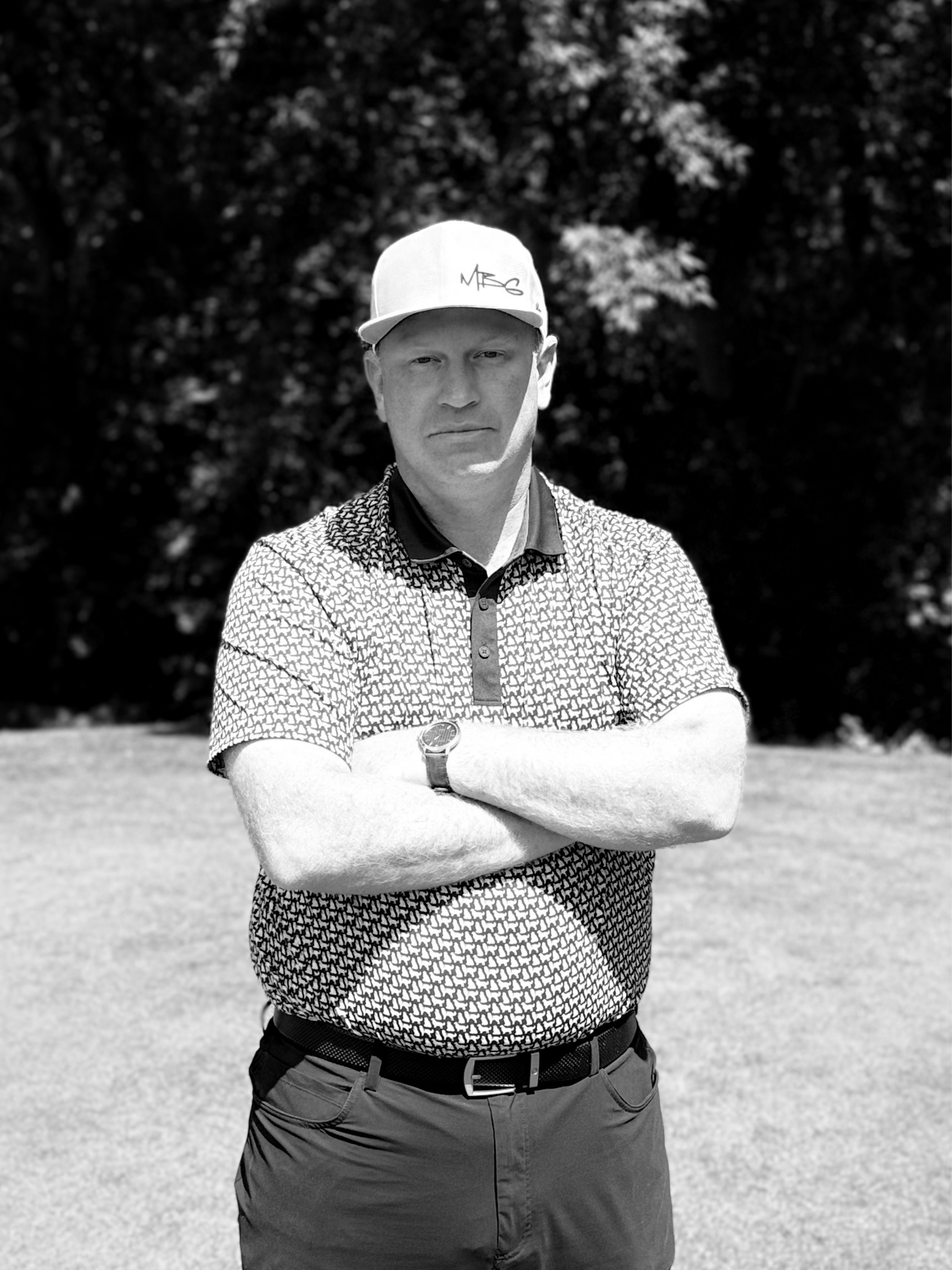 A young man standing outdoors with arms crossed, wearing a patterned golf shirt, dark pants, a baseball cap, and a watch, with trees in the background.