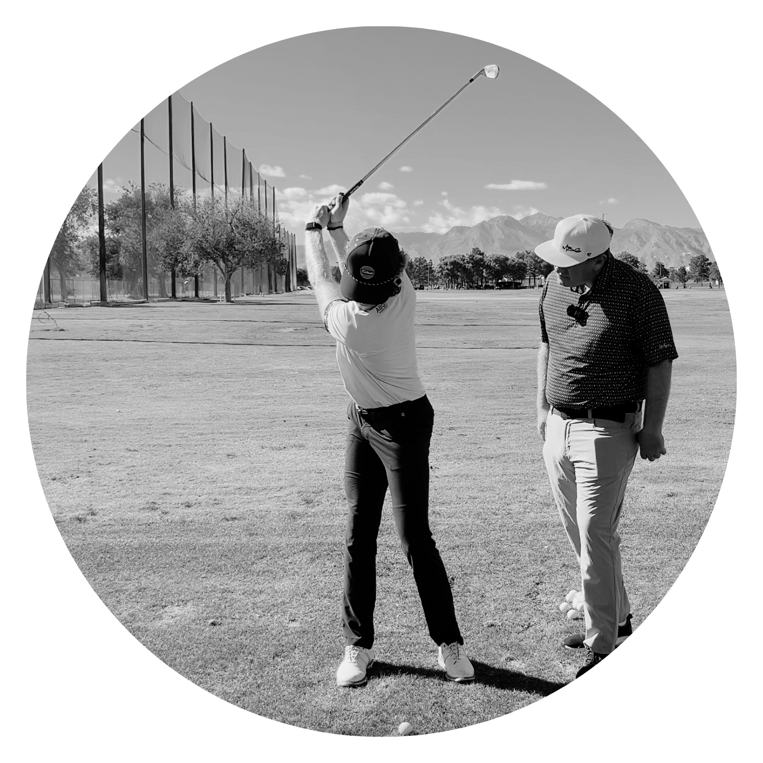 A person holding a golf club preparing to hit a golf ball on a golf course with another person watching, mountains in the background, and trees along the field.