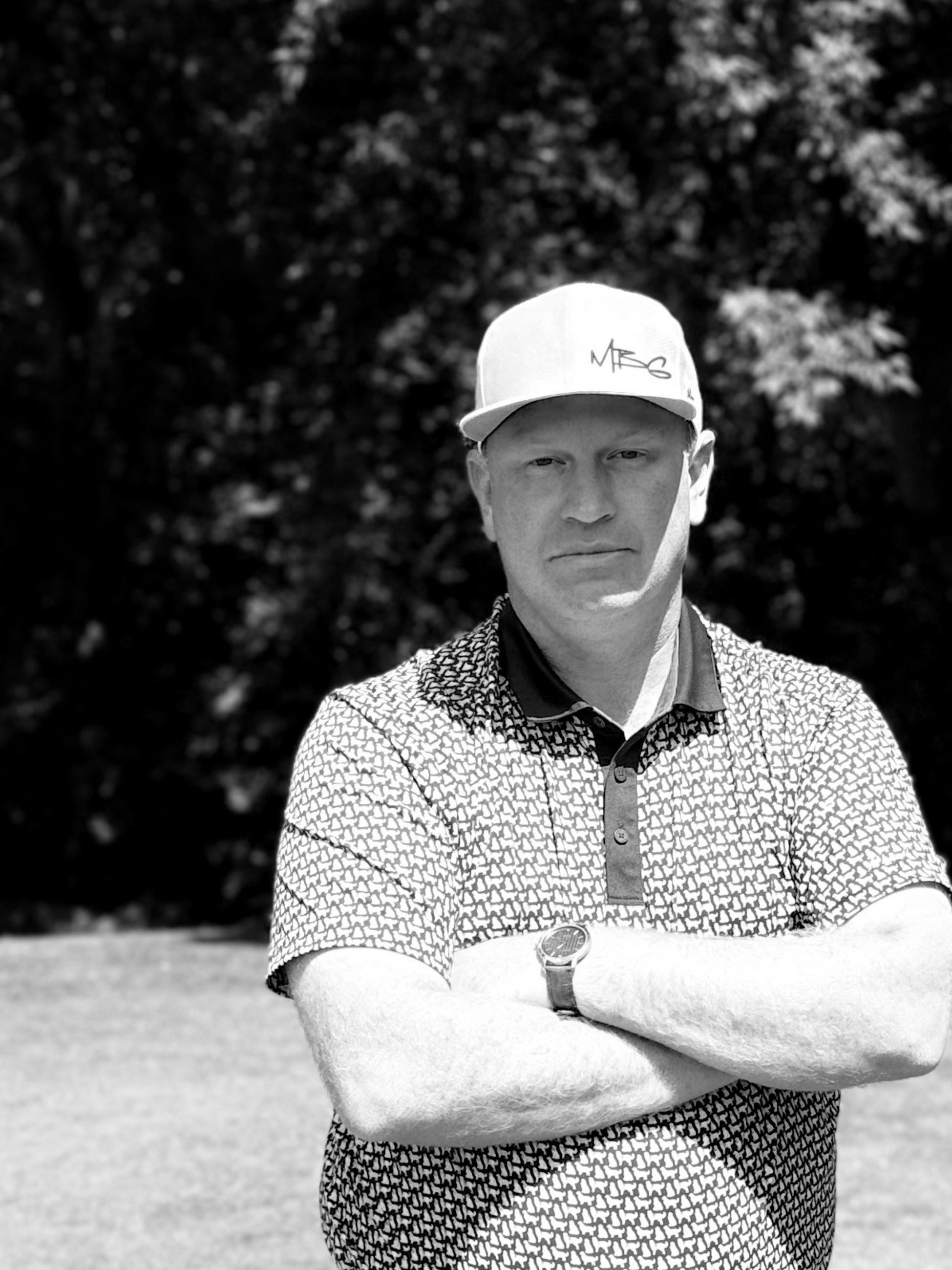 A man wearing a baseball cap and patterned polo shirt, standing outdoors with trees in the background.