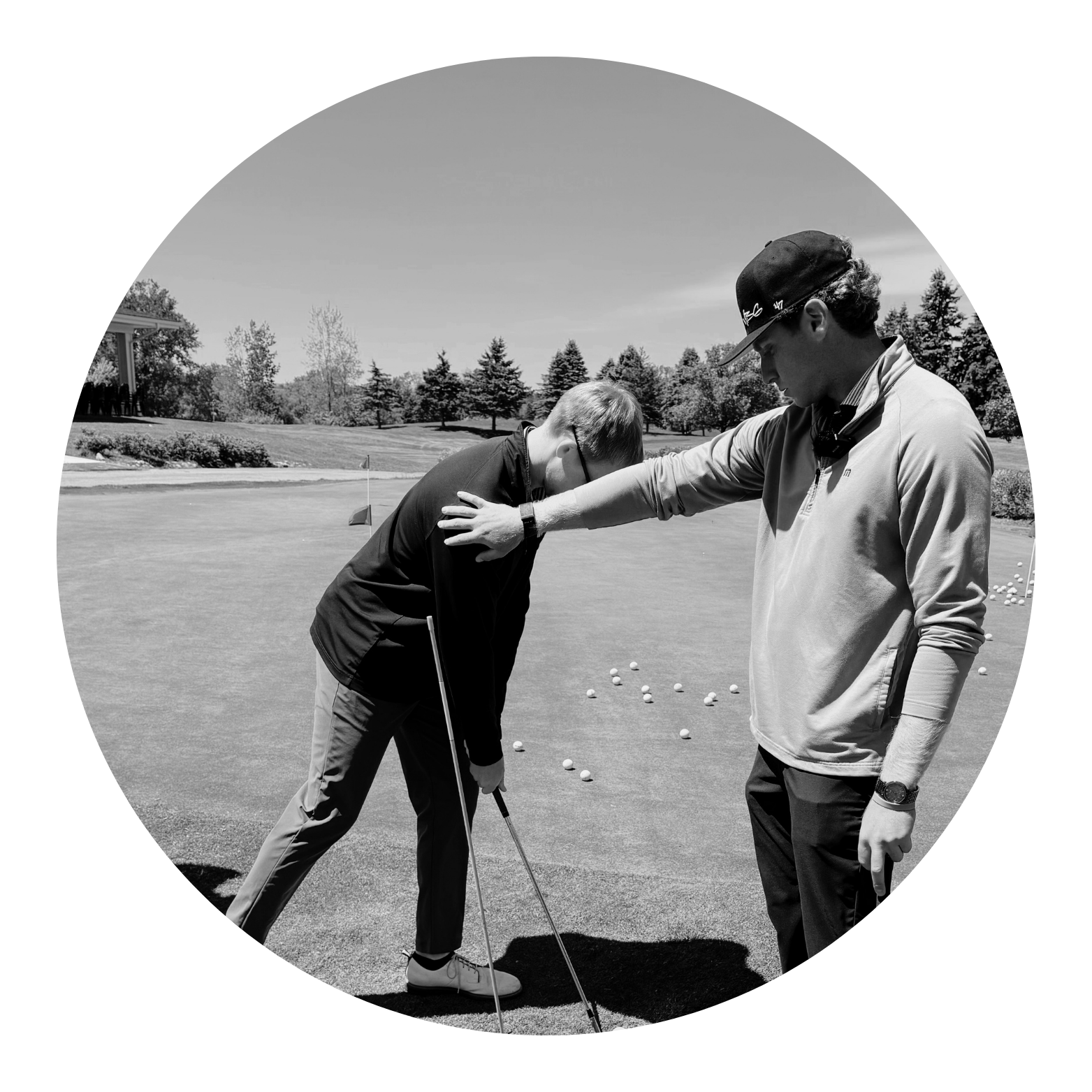 Two men are on a golf course, with the younger man leaning over to position a golf ball. The older man, wearing a cap and a long-sleeved shirt, is giving instructions. Multiple golf balls are scattered around them on the putting green, with trees and a partly cloudy sky in the background.