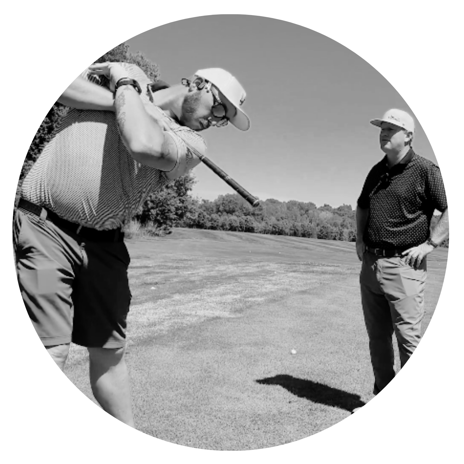 Two men are on a golf course, one is preparing to hit a golf ball with a club while the other observes. They are wearing hats and casual golf attire, with trees in the background under a clear sky.