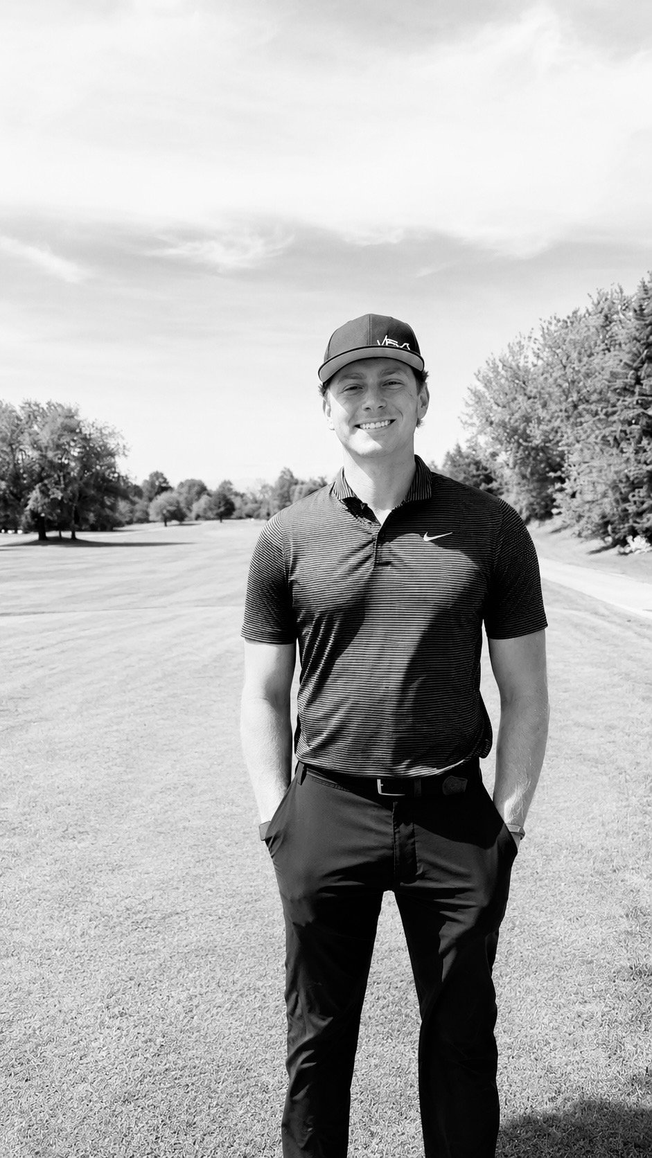 A young man standing on a golf course with trees in the background, wearing a baseball cap, a striped Nike polo shirt, and dark pants, smiling at the camera.