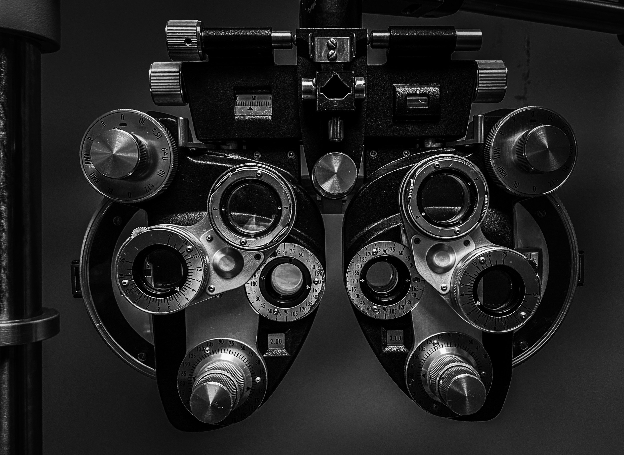 Black and white close-up image of a phoropter, an eye examination device with various dials and lenses.