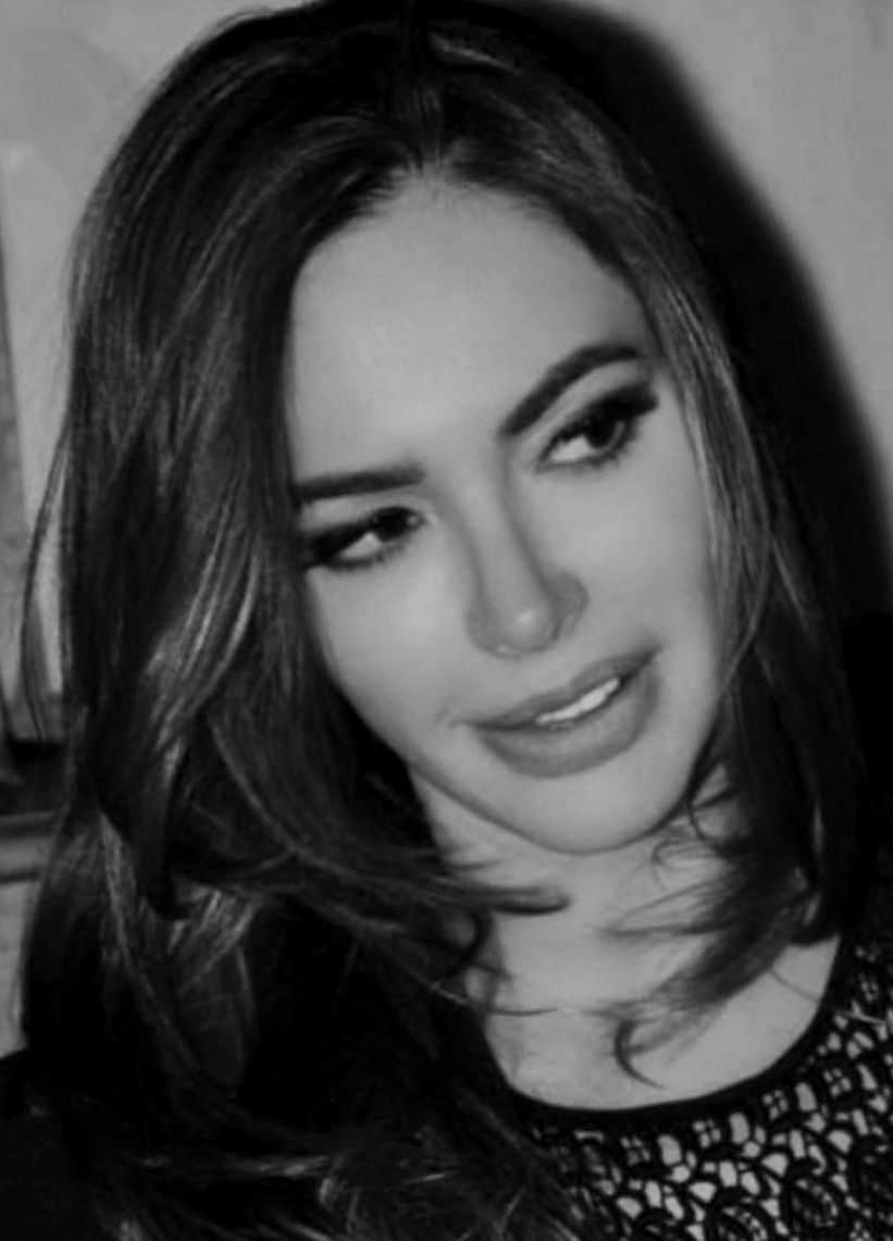 A young woman with long dark hair, styled in loose waves, looking to her left with a slight smile. She has dark eyebrows and makeup, and is wearing a patterned top. The photo is in black and white.