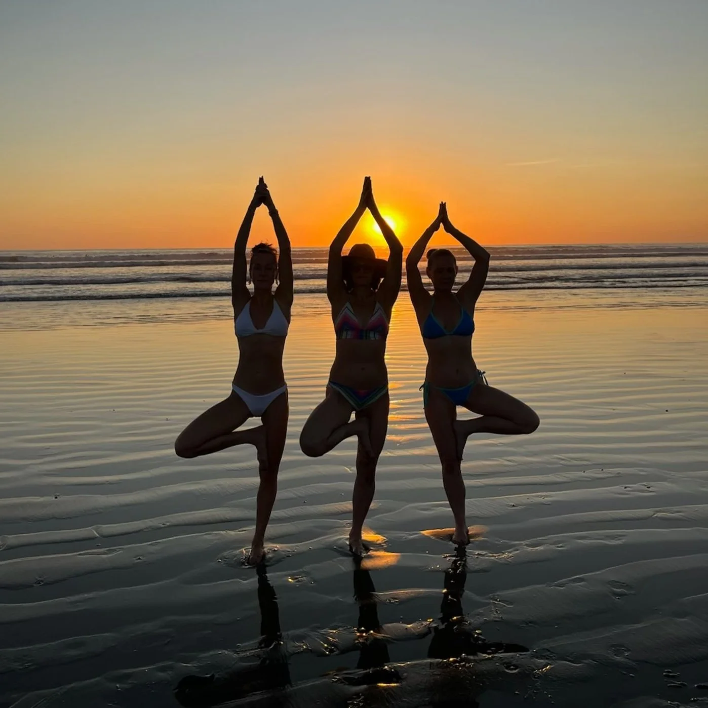 Three women practicing yoga on the beach at sunset, standing on one leg in tree pose with hands pressed together overhead.