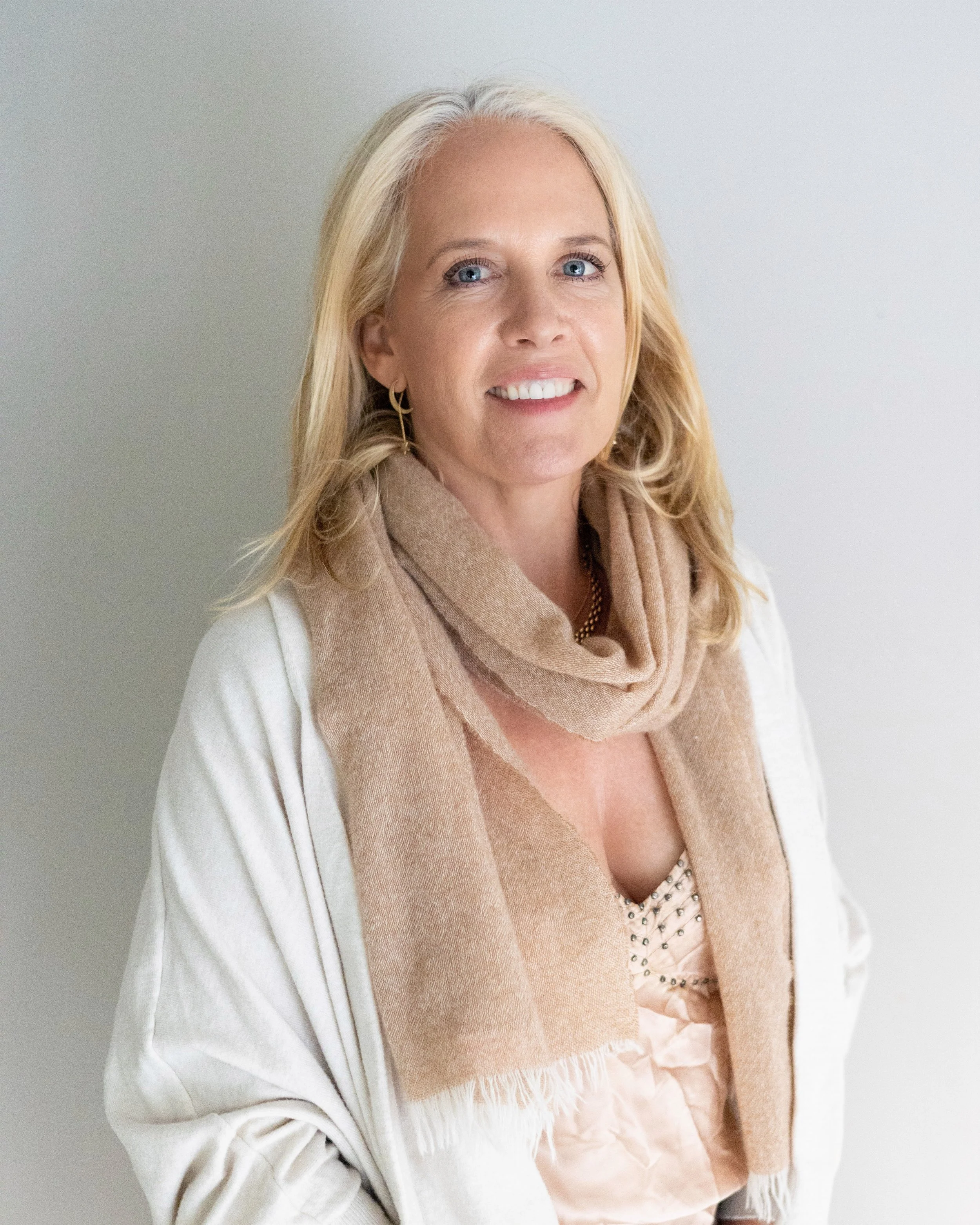 A middle-aged woman with blonde hair, blue eyes, and earrings, smiling, wearing a beige scarf and light-colored clothing, standing against a light background.