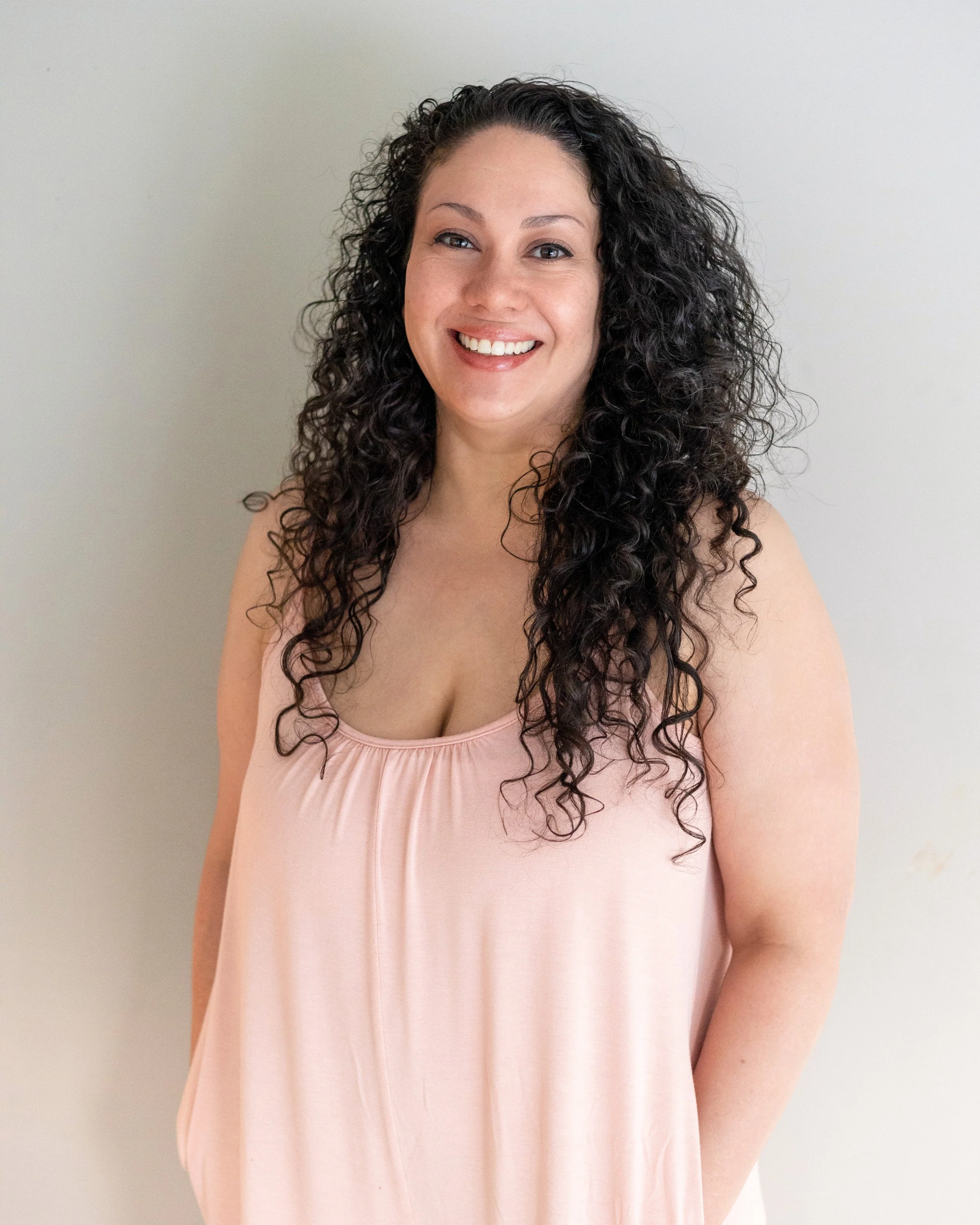 A smiling woman with long curly dark hair wearing a light pink sleeveless top standing against a plain white wall.