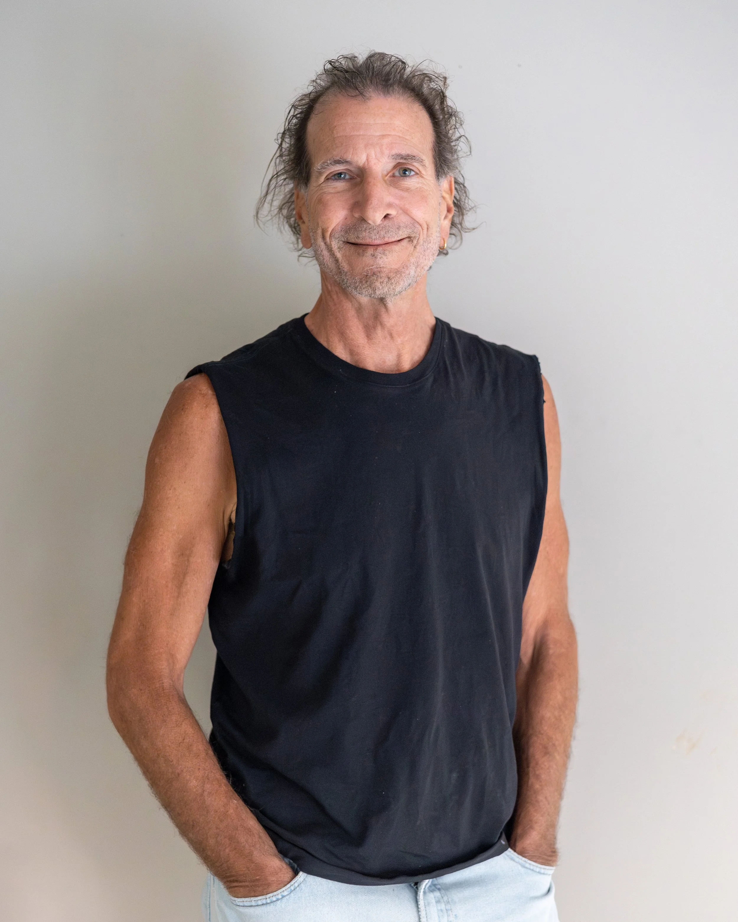 A middle-aged man with curly hair, wearing a black sleeveless shirt and light pants, standing against a plain white wall, smiling and looking at the camera.