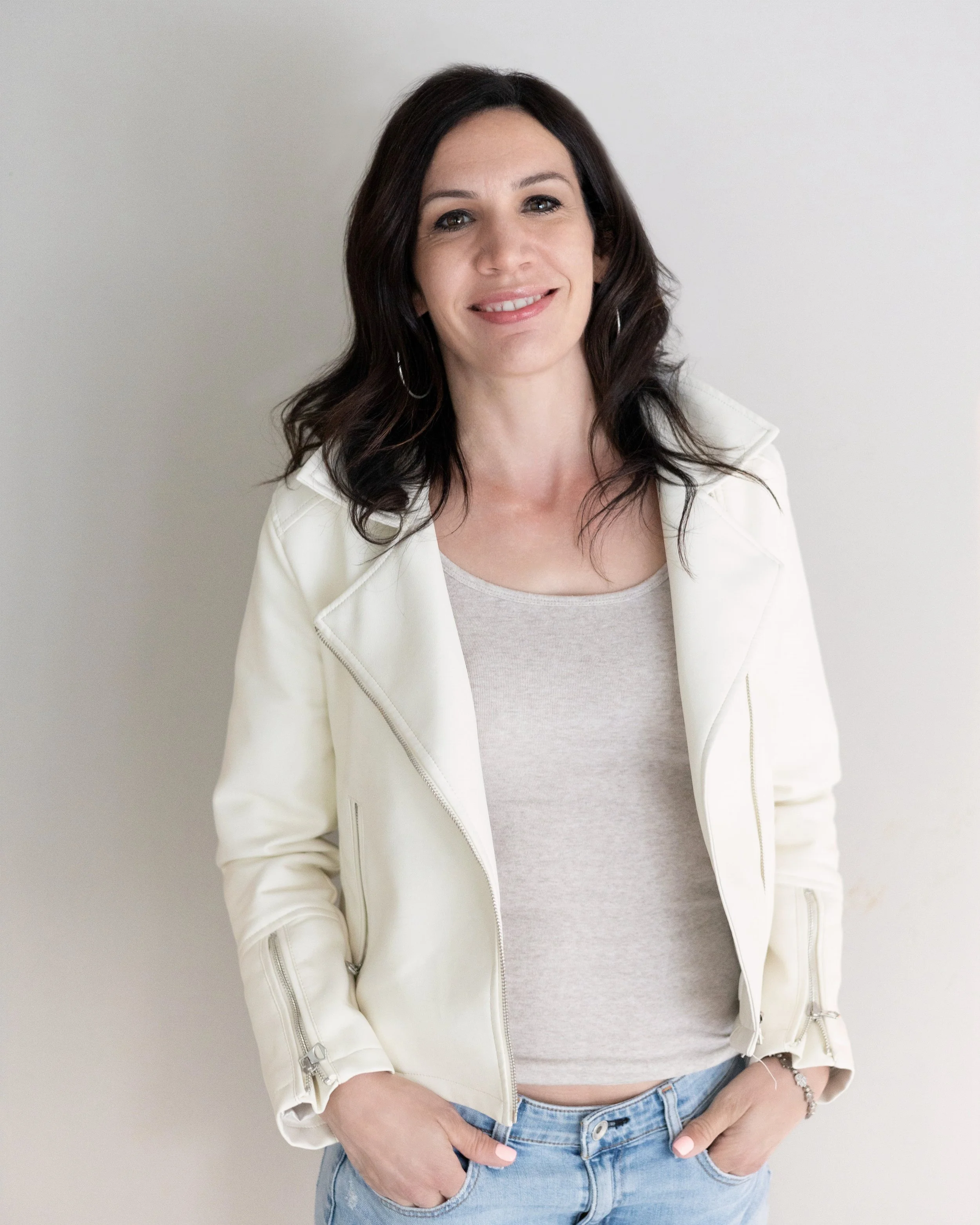 Smiling woman with dark wavy hair wearing a white leather jacket, gray top, and blue jeans, standing against a plain light-colored background.