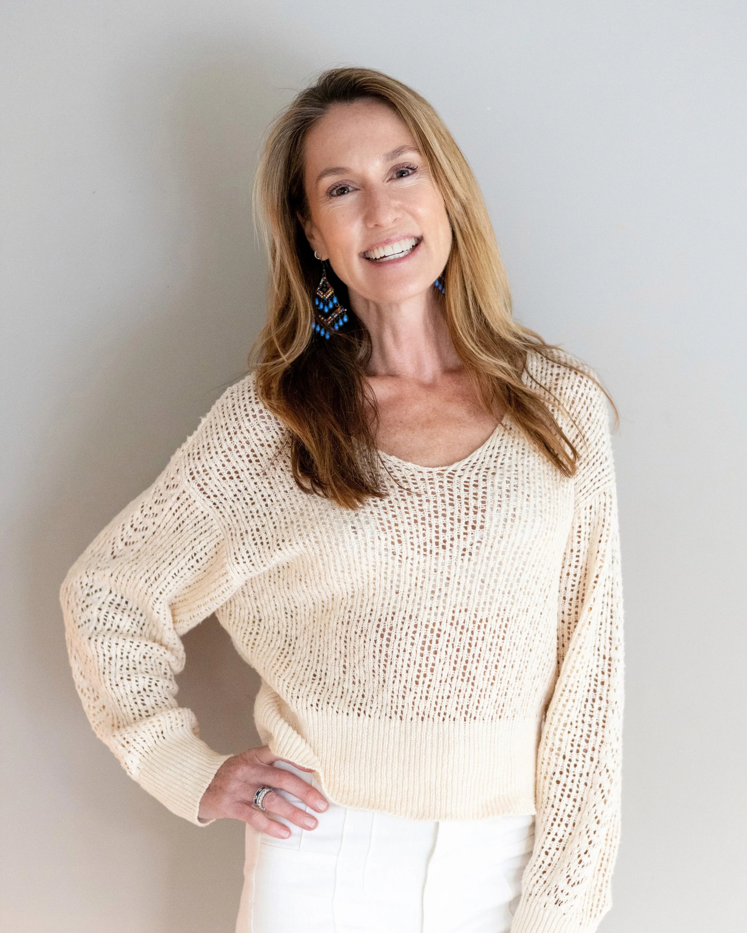 A smiling woman with long, wavy, light brown hair wearing a cream-colored crochet sweater and white pants, standing against a plain light-colored wall.