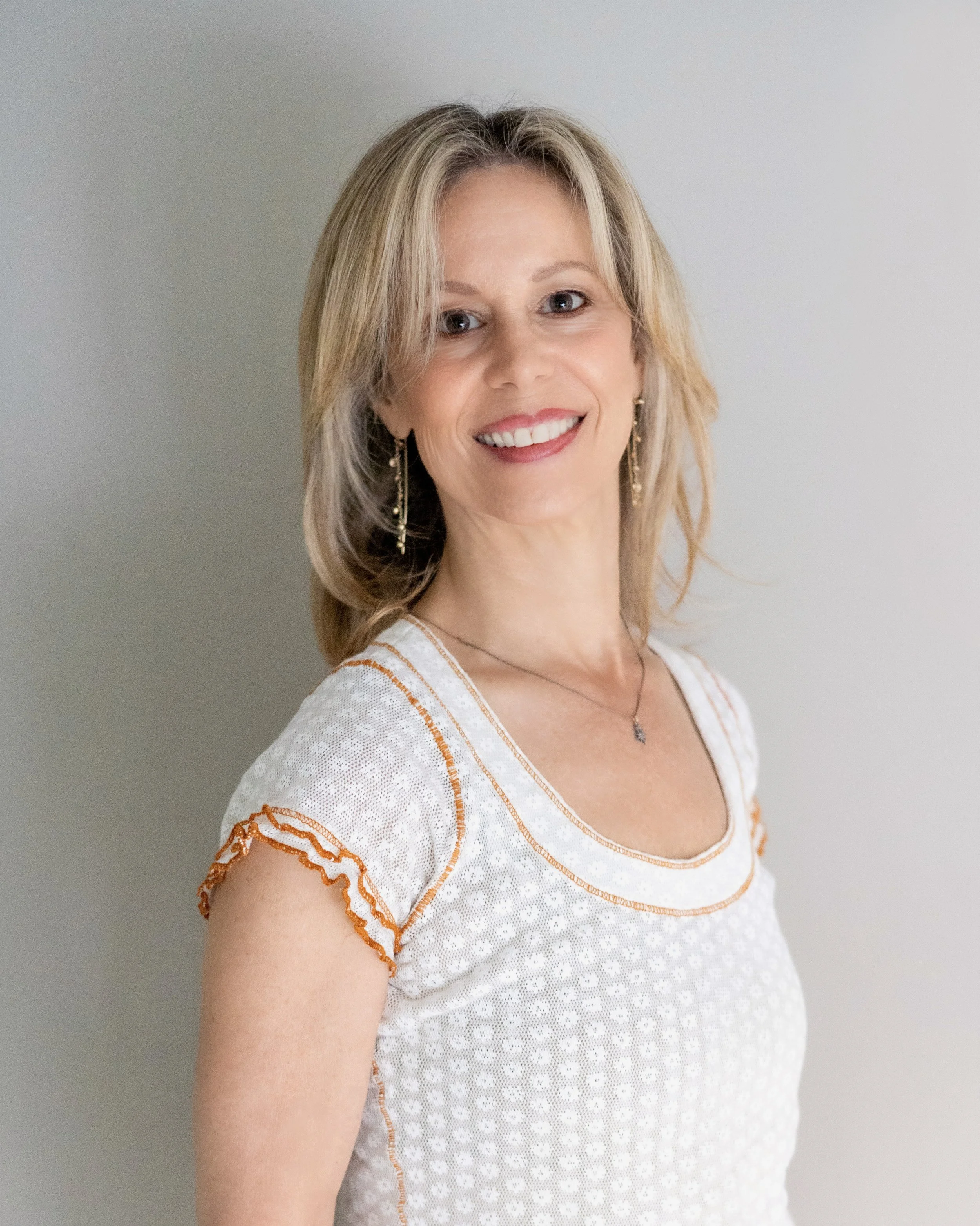 A woman with shoulder-length blonde hair is smiling at the camera. She is wearing a white top with orange and beige accents, gold earrings, a necklace, and is standing against a plain white background.