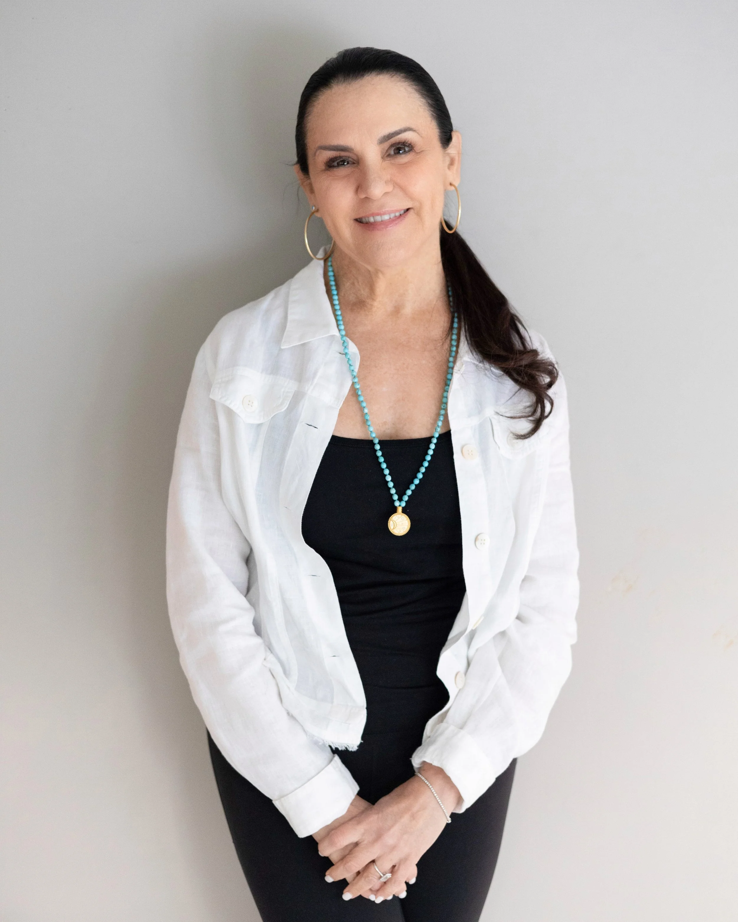 A smiling woman with dark hair pulled back, wearing a black top, white jacket, gold hoop earrings, turquoise necklace with a gold pendant, and a silver bracelet, standing against a plain light-colored wall.