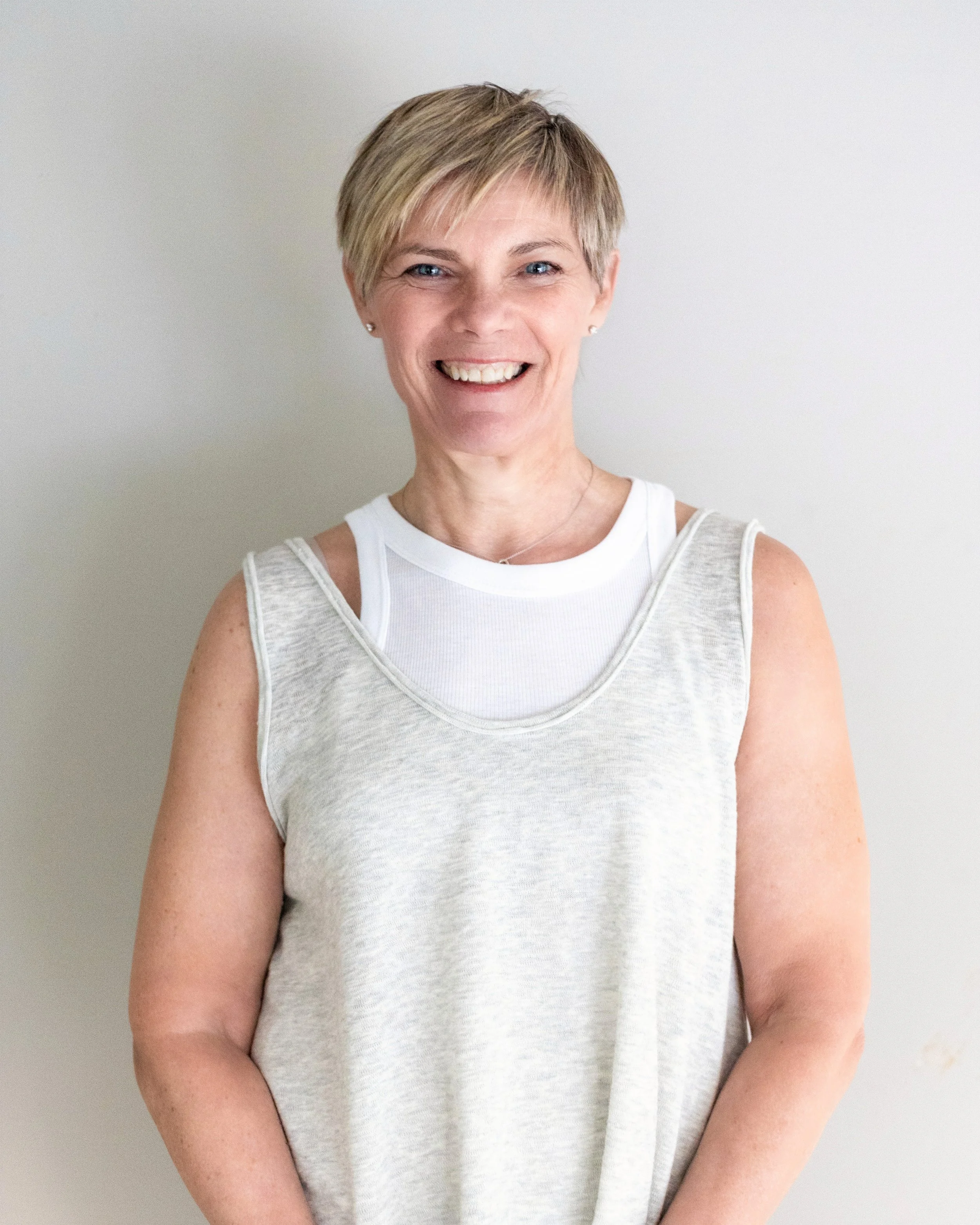 A woman with short blonde hair smiling, wearing a white sleeveless top and a white undershirt, standing against a plain white wall.