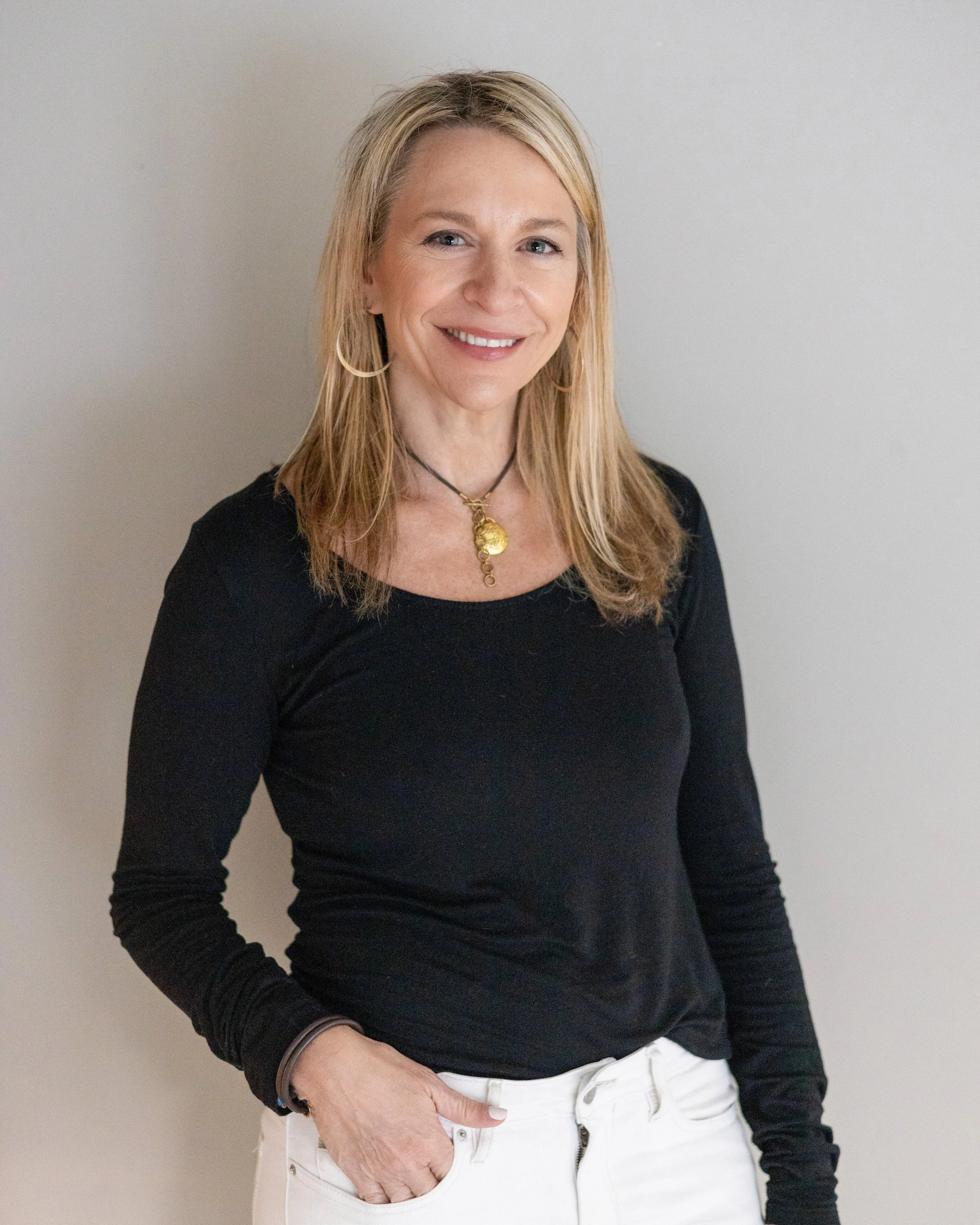 A woman with blonde hair, wearing a black long-sleeve shirt, white pants, hoop earrings, and a necklace, standing against a plain light-colored wall and smiling.