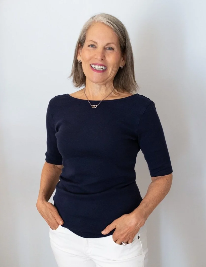 A smiling middle-aged woman with gray hair, wearing a navy blue top, white pants, and a necklace with a small infinity symbol pendant, standing against a plain light background.