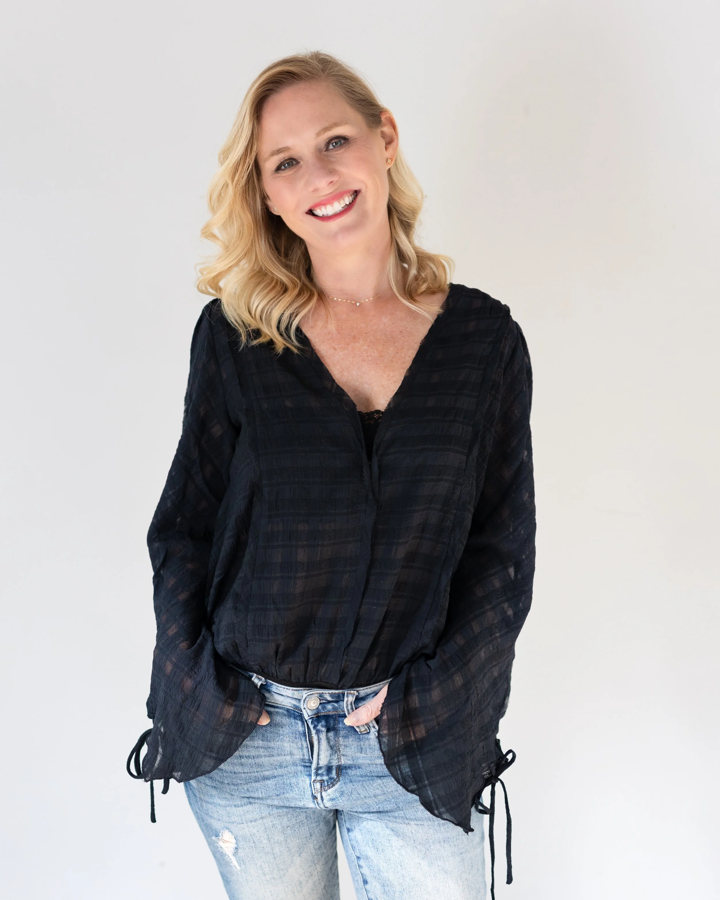 A woman with shoulder-length blond wavy hair, smiling, wearing a black long-sleeve top with a subtle checkered pattern and light blue ripped jeans, standing against a plain white background.