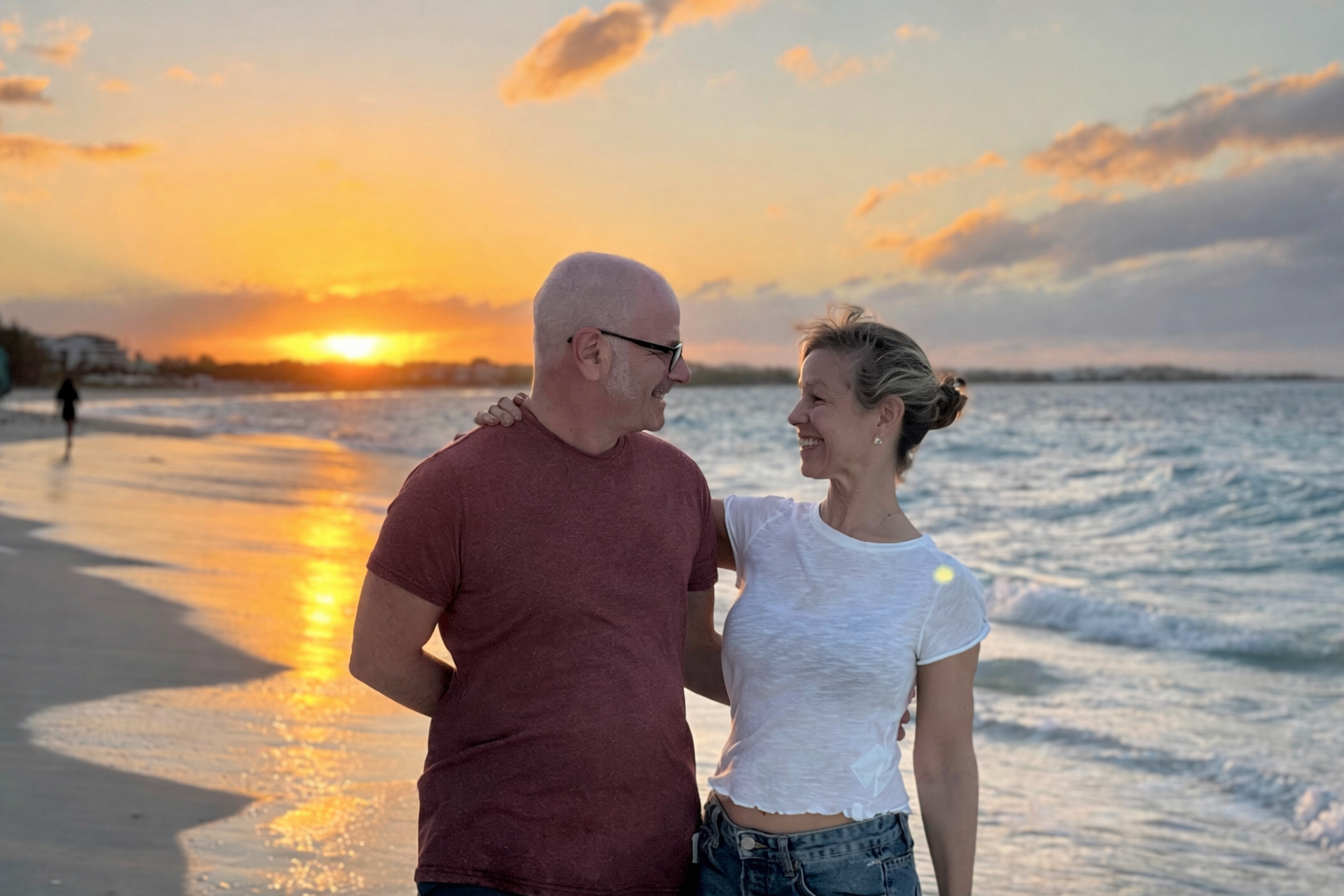 An older man and woman smiling and looking at each other, standing on the beach during sunset.