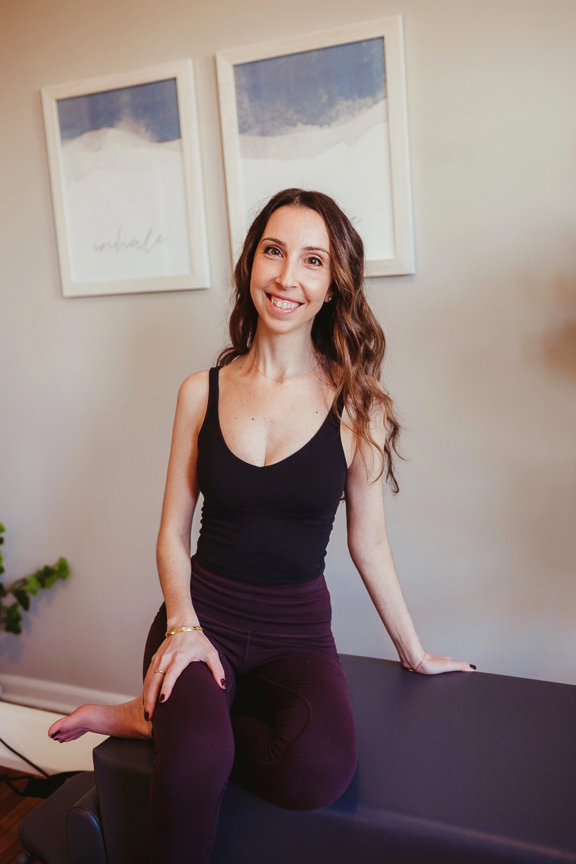 Woman with long brown hair smiling in a black tank top and dark purple leggings, sitting on a black massage table in a room with neutral decor and framed artwork on the wall.