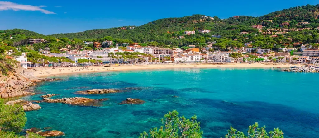 Coastal town with white buildings along a sandy beach, turquoise water, and green hills in the background under a clear blue sky.