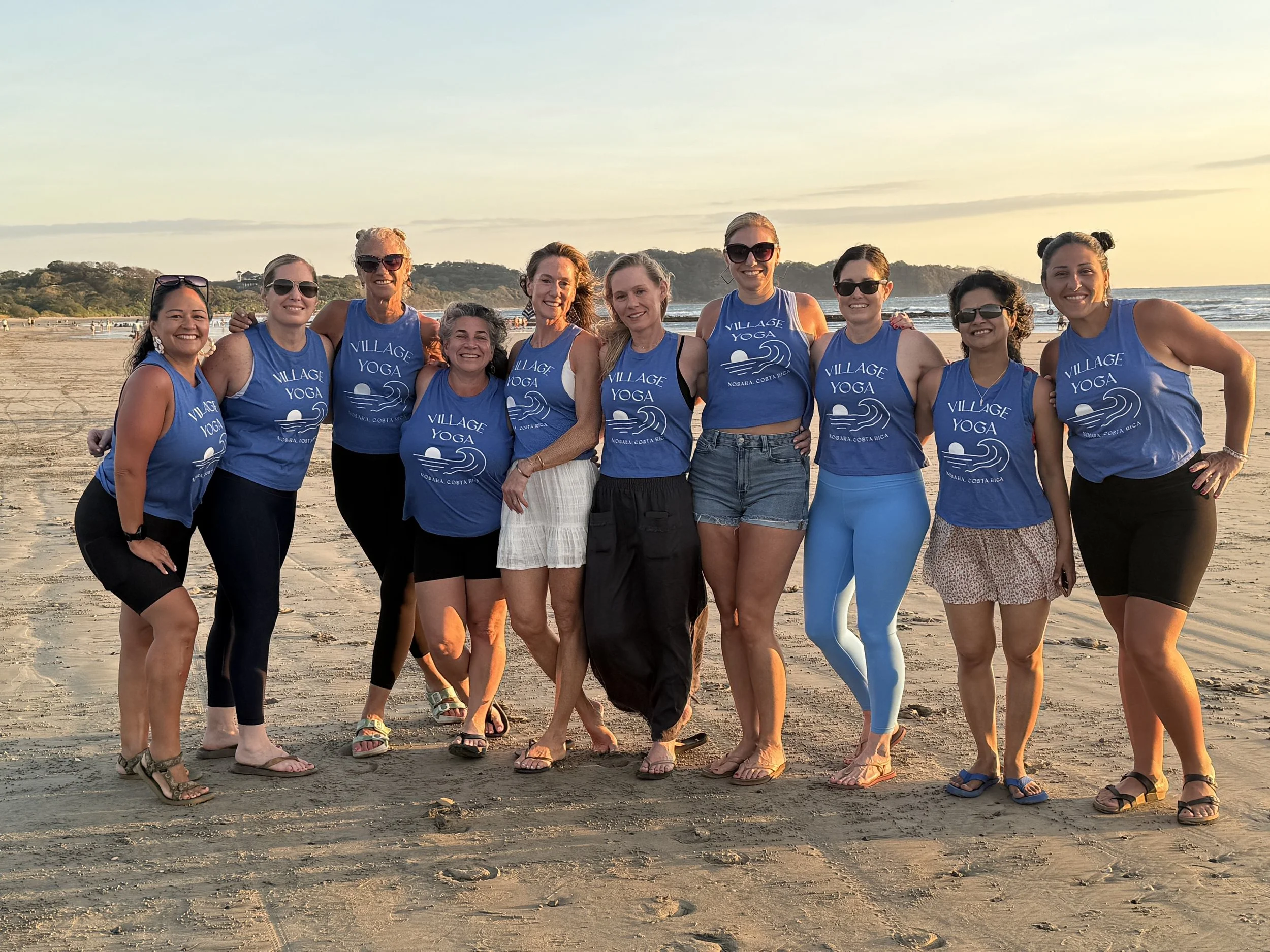 Group of women on the beach during sunset, wearing matching blue village yoga shirts, smiling and posing for the photo.