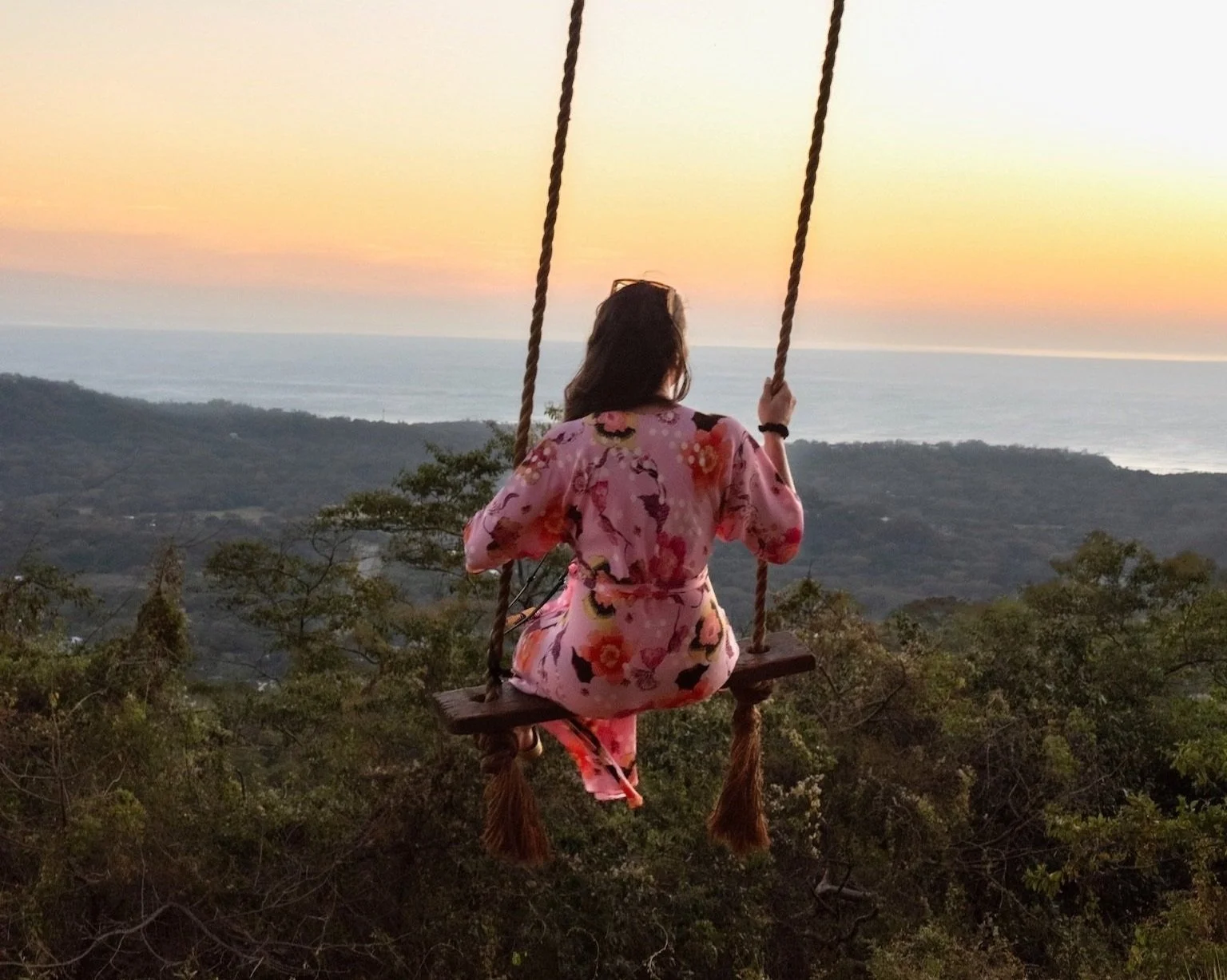 A woman in a pink floral dress sitting on a swing overlooking a landscape with trees, hills, and an ocean at sunset.