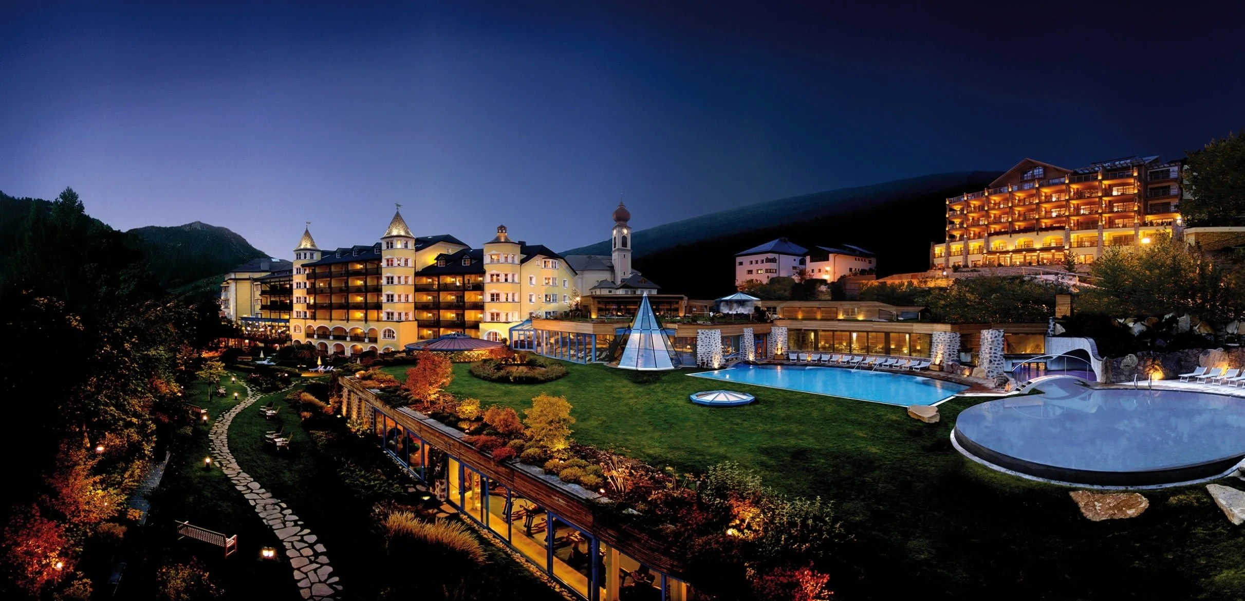 A resort at night with illuminated buildings, a swimming pool, and mountain scenery in the background.