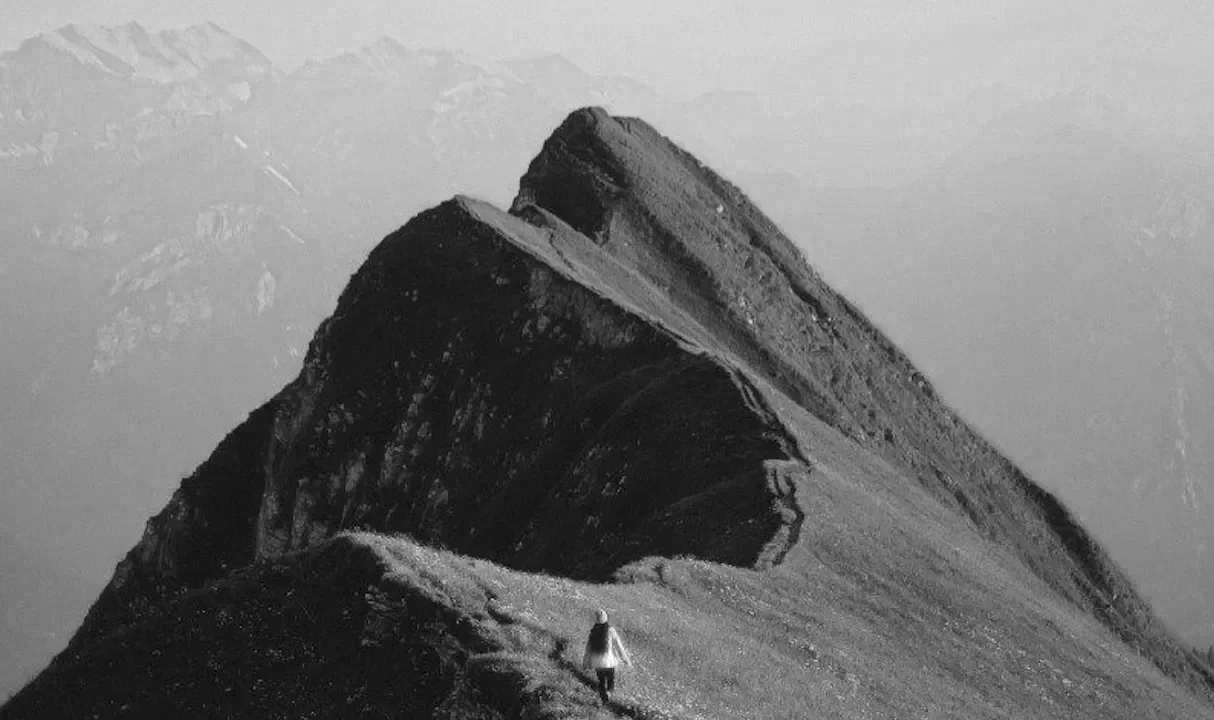 A person walking on a winding mountain trail with a mountain peak in the background.