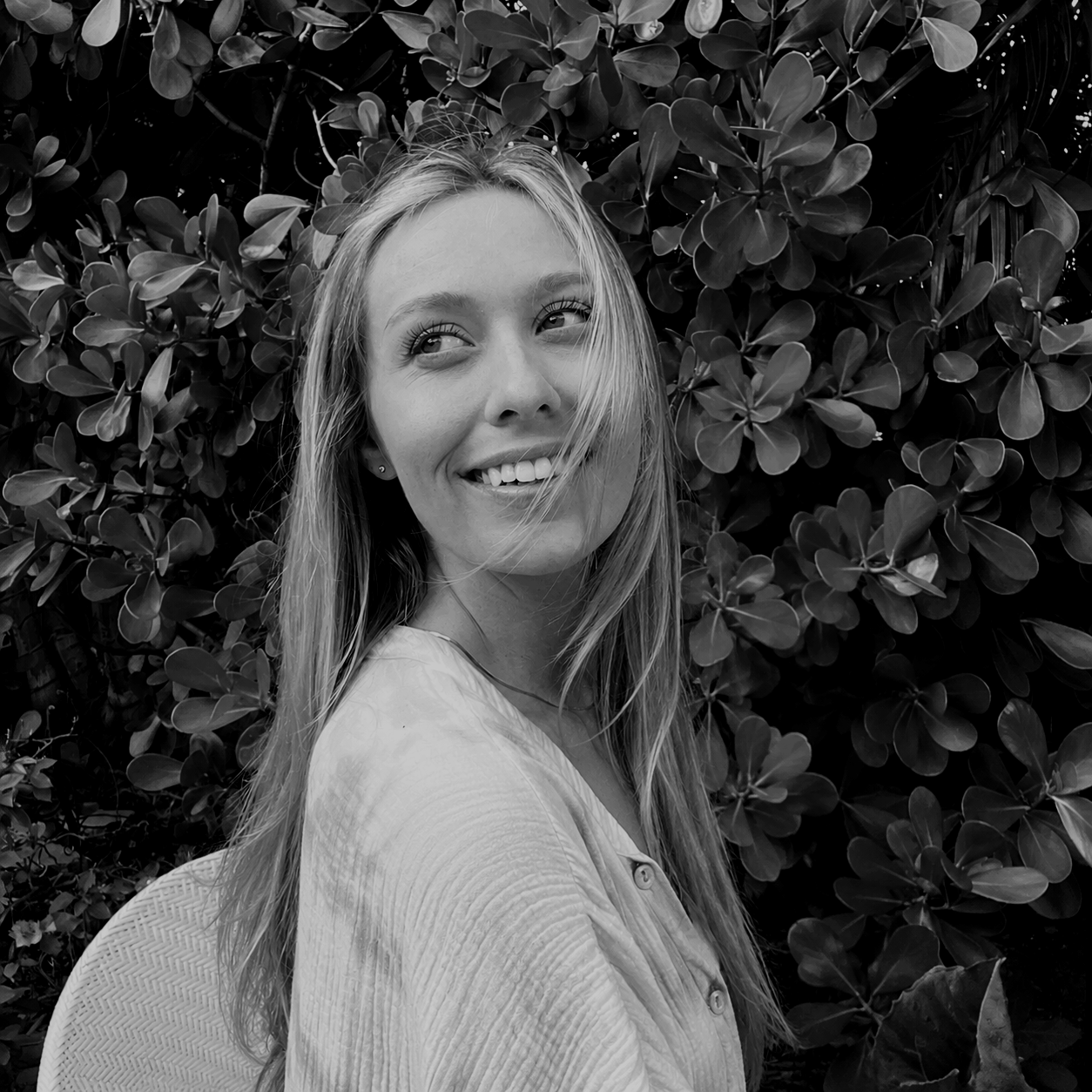 Black and white photo of a smiling woman with long hair sitting outdoors in front of a leafy bush, looking slightly to the side.