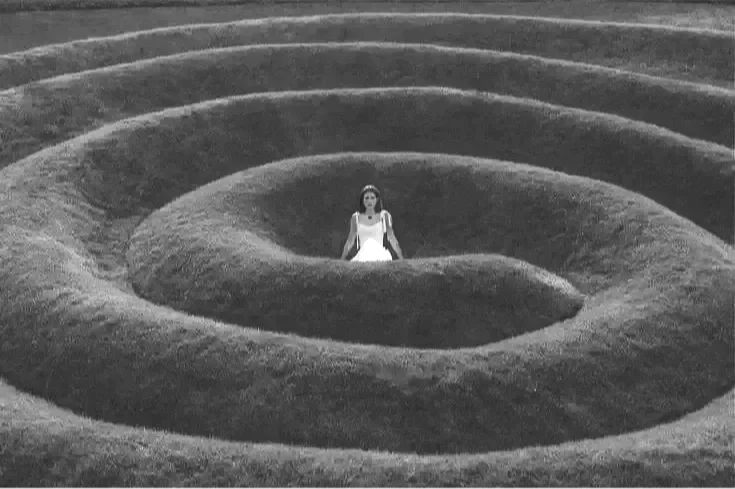 A woman in a white dress stands in the center of a spiral labyrinth made of grass.