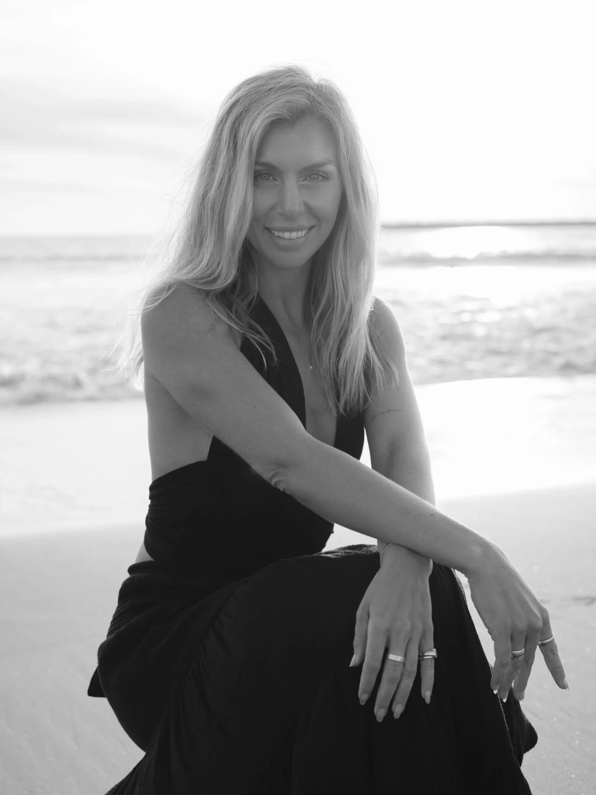 Black and white photograph of a smiling woman with long hair sitting on a beach, with the ocean in the background.