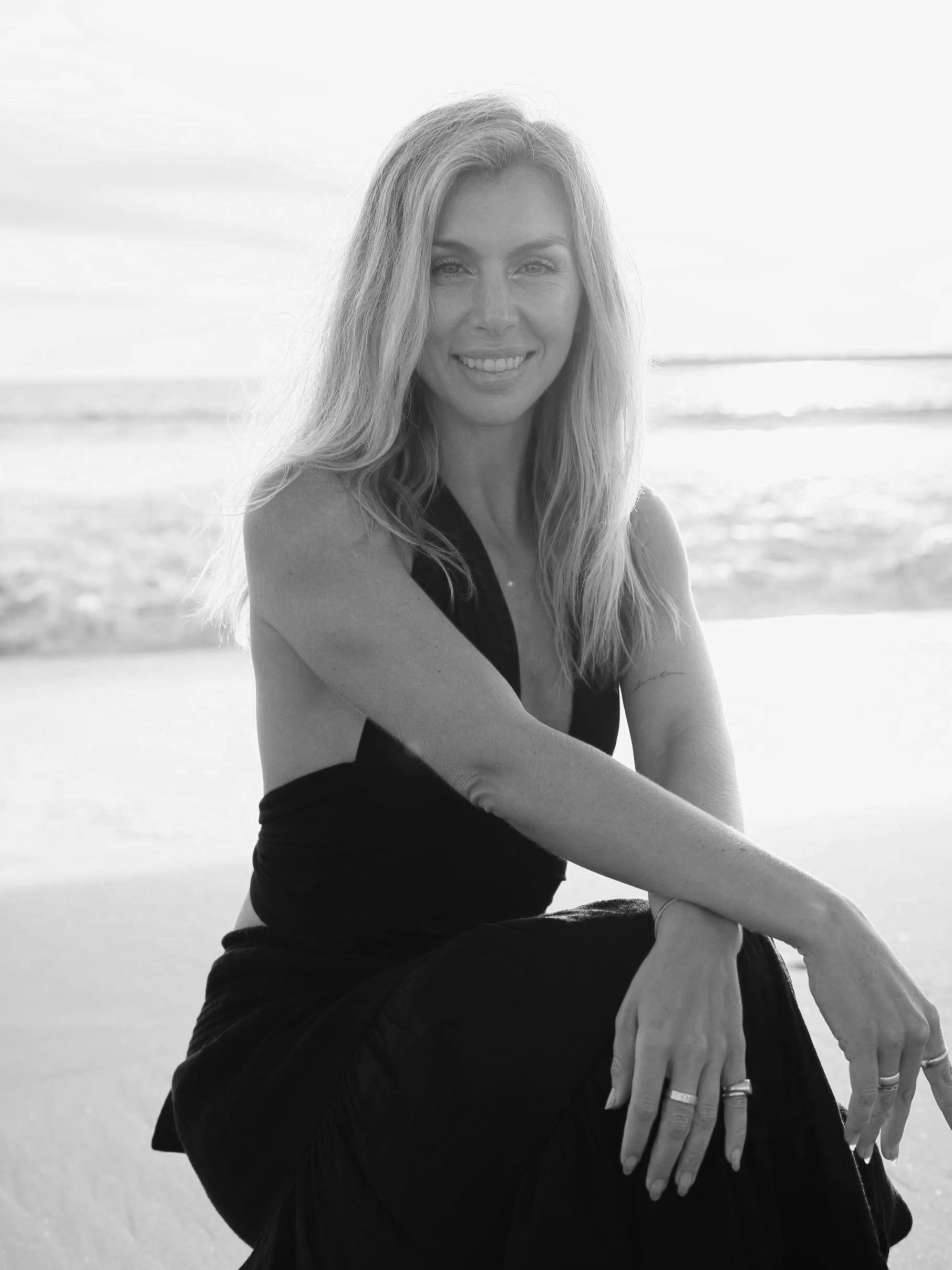 Black and white photo of a woman sitting on the beach, smiling, with the ocean and sky in the background.
