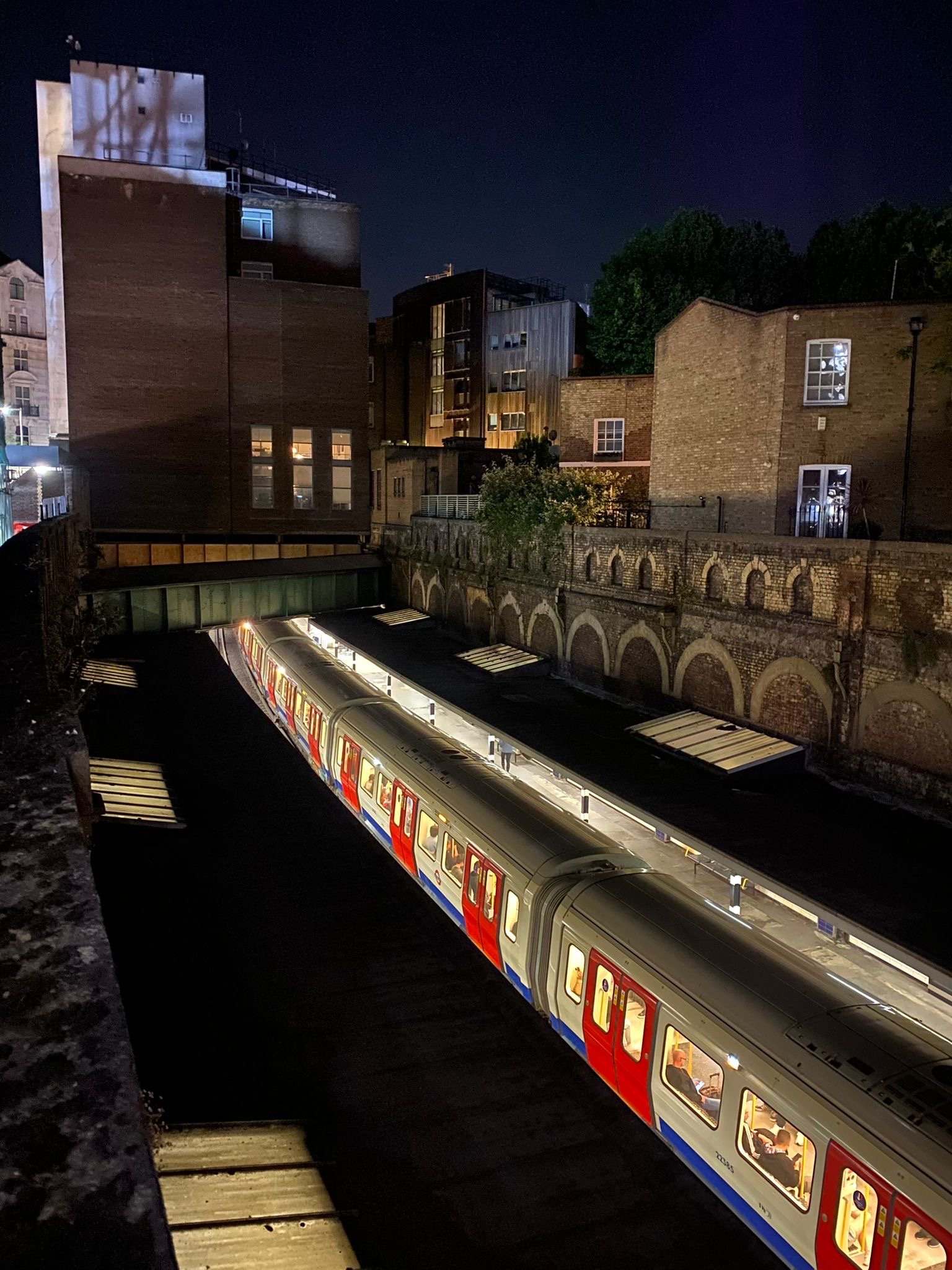 Nighttime scene of a London Underground train at a station, with surrounding buildings and trees in the background.