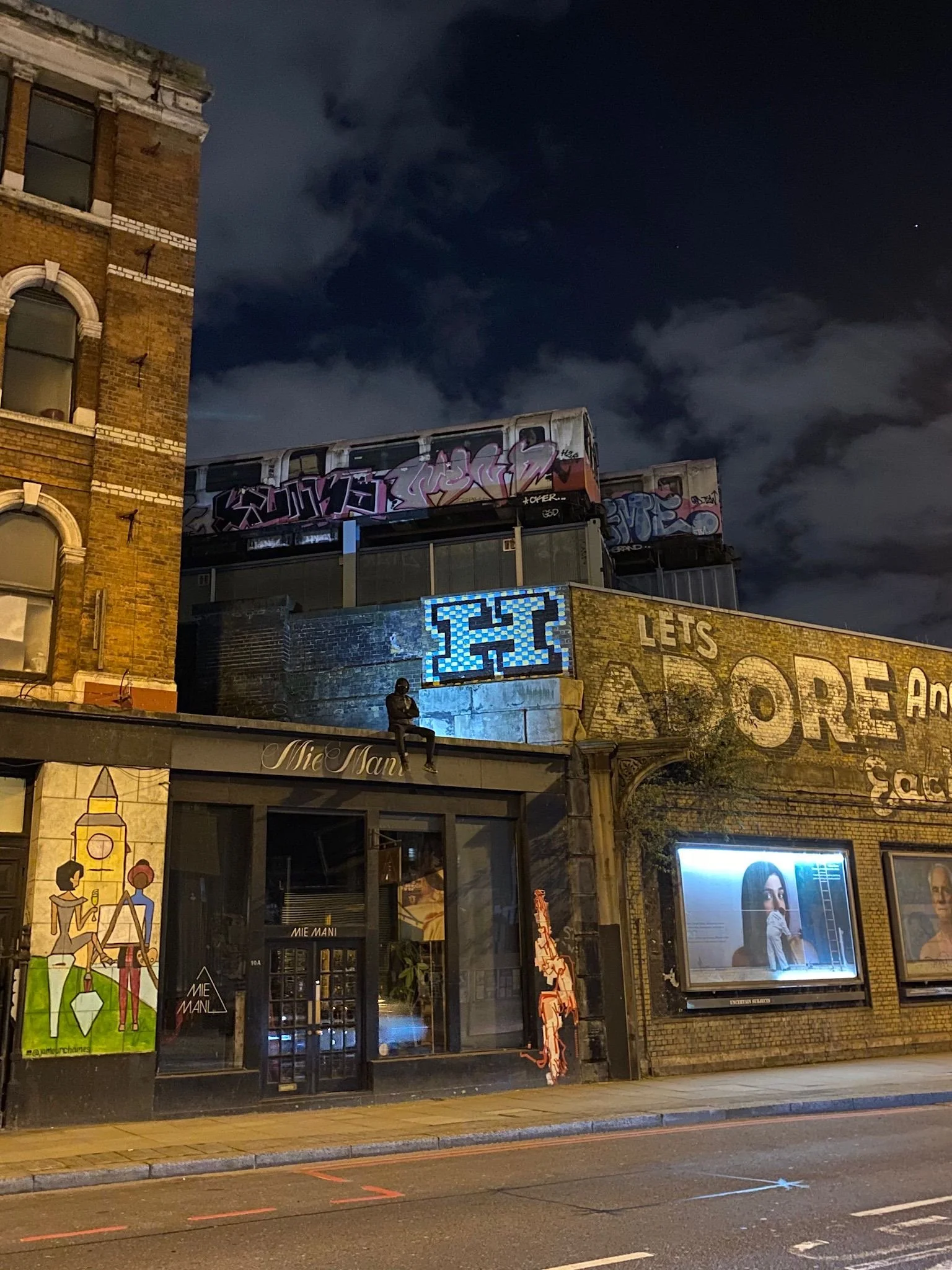 Nighttime urban scene showing a brick building with painted artwork, graffiti, and a building with illuminated signs. A person is sitting on a ledge outside a store.