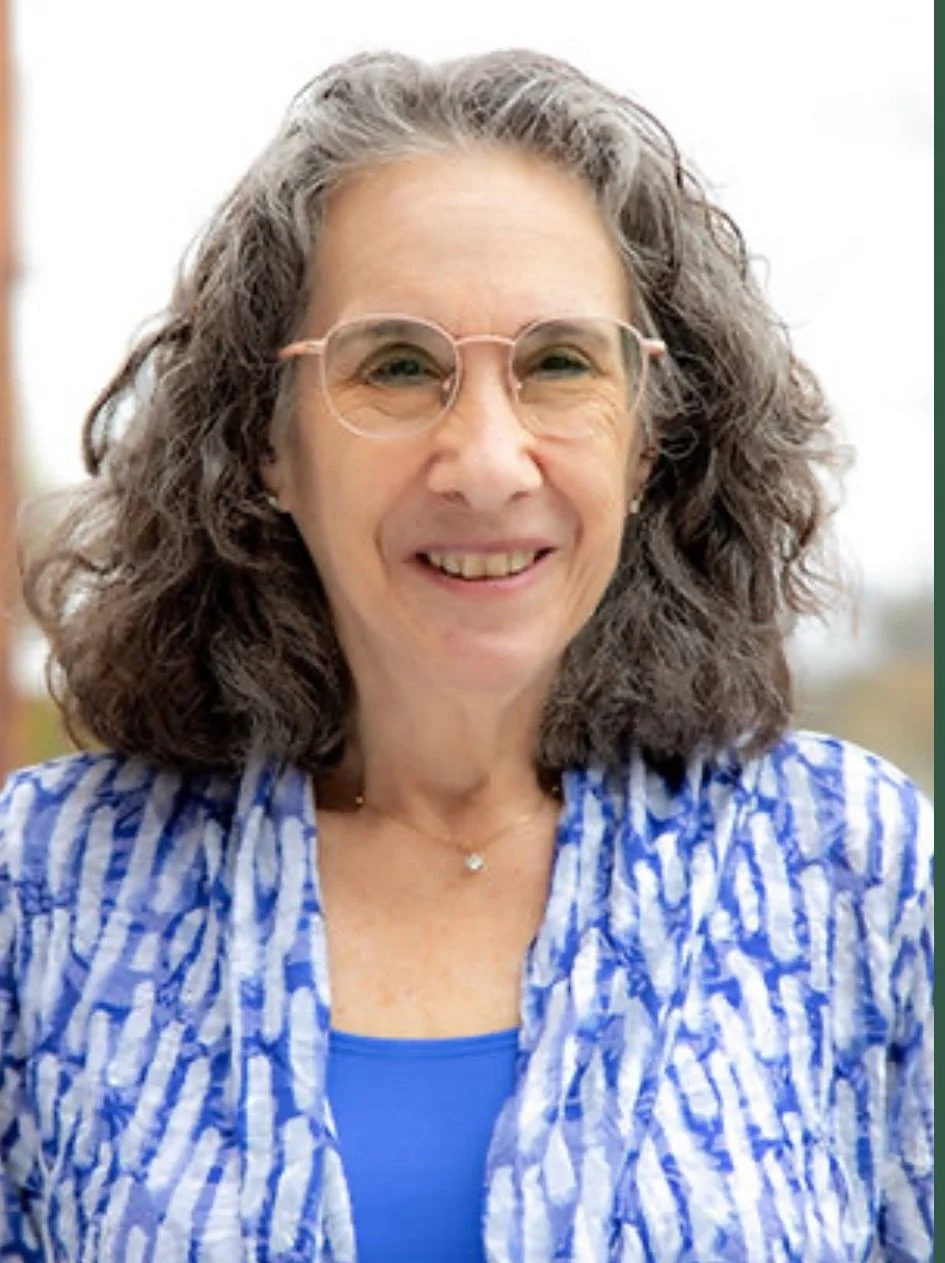 A middle-aged woman with curly gray hair, wearing glasses, a blue patterned shirt, and a blue top underneath, smiling outdoors.