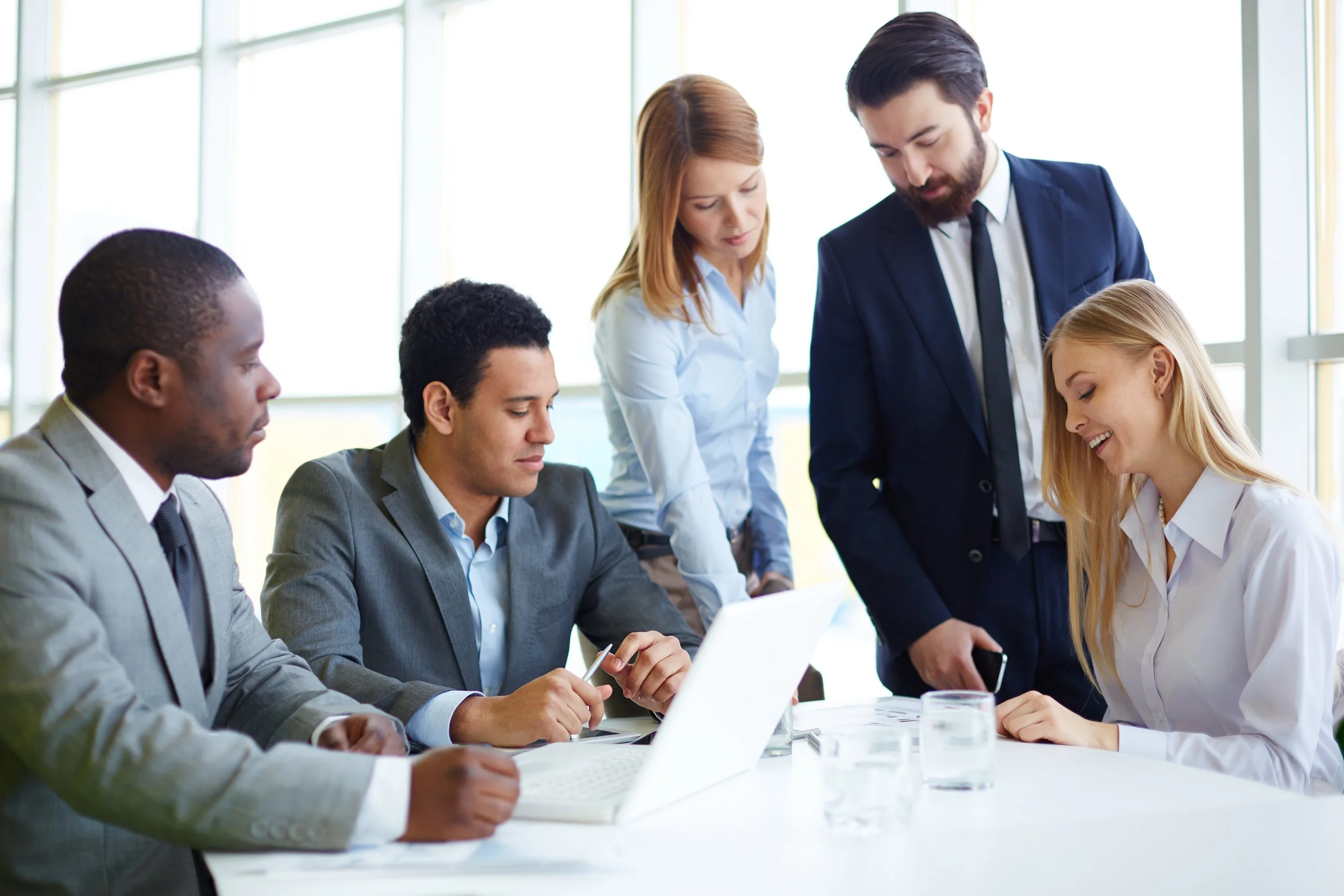 Business meeting with five professionals, three men and two women, gathered around a table in a bright office, reviewing documents and discussing.
