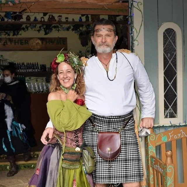 A smiling woman and a man in traditional Scottish attire, standing indoors. The woman wears a floral headpiece and colorful skirt, while the man wears a kilt and a white shirt.