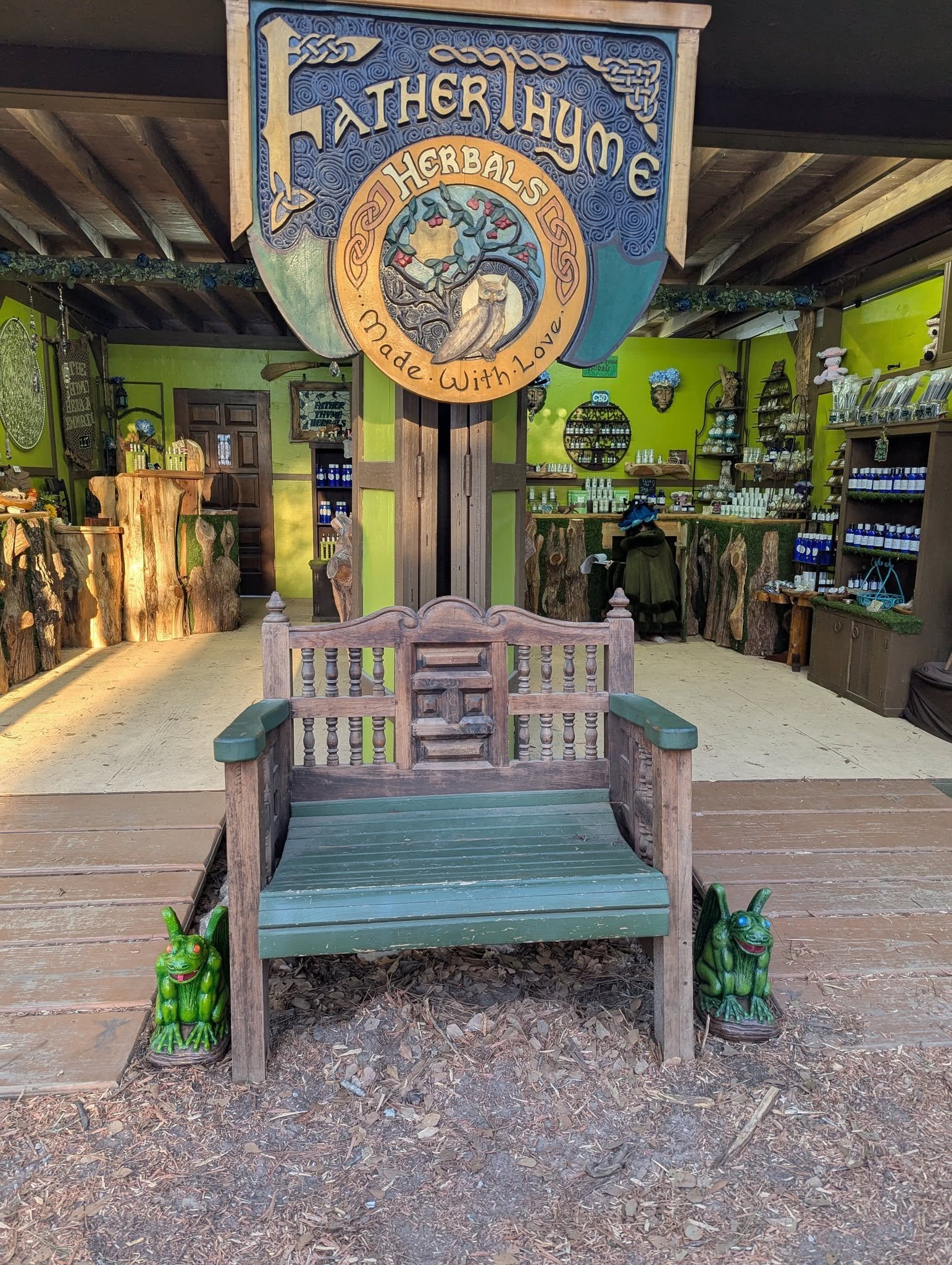 Outdoor scene featuring a bench with foliage at the base, surrounded by two green frog statues on either side. In the background, there are shelves with various bottles and products, a wooden door, and a sign hanging from the ceiling that reads 'Father Thyme Herbal's' with a decorative owl and tree design, all under a wooden roof structure.
